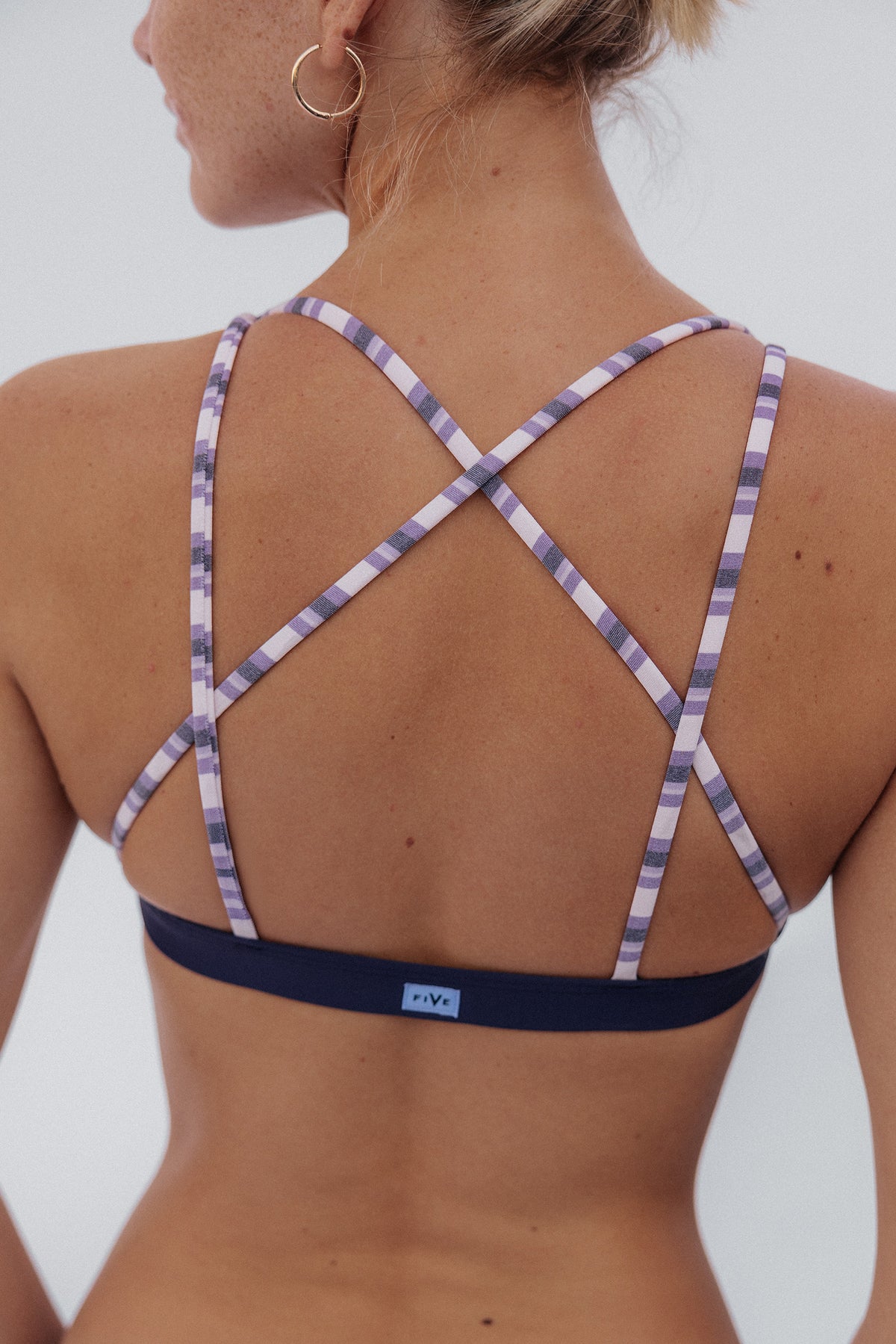 women, Close-up of a woman wearing a purple sports bra with purple and white striped straps and underband, shown against a light gray background.