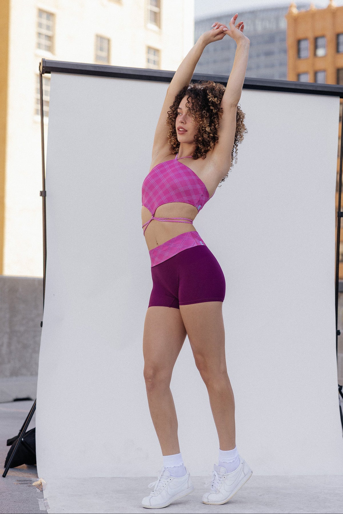 women, posing in a pink and purple athletic set consisting of a crop top and shorts, standing in front of a white studio backdrop with city buildings in the background.