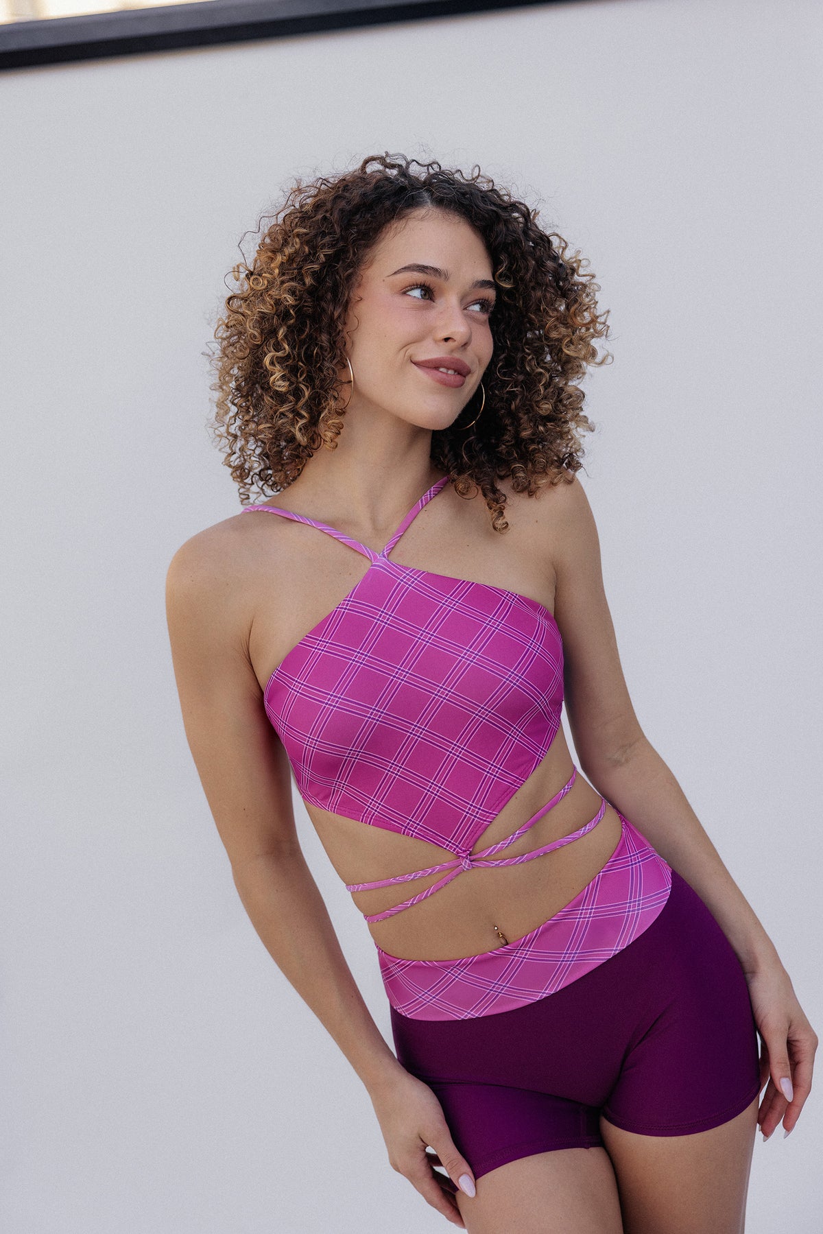 women, modeling a two-piece activewear set featuring a pink and white plaid halter crop top with side cutouts and matching purple athletic shorts, set against a plain light gray background.