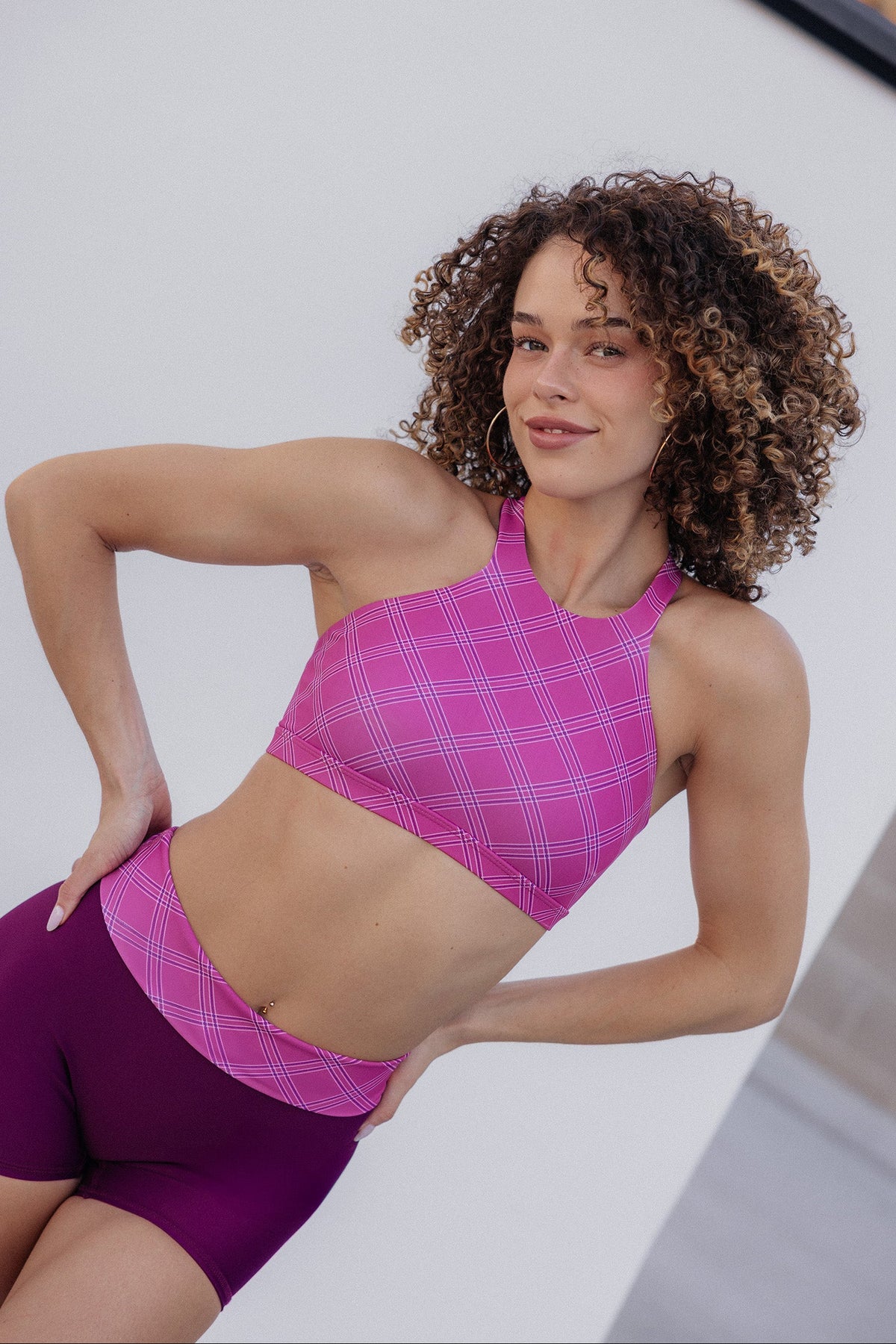 women, with curly brown hair wearing a pink and purple plaid sports bra and matching high-waisted leggings, posing against a light gray background.