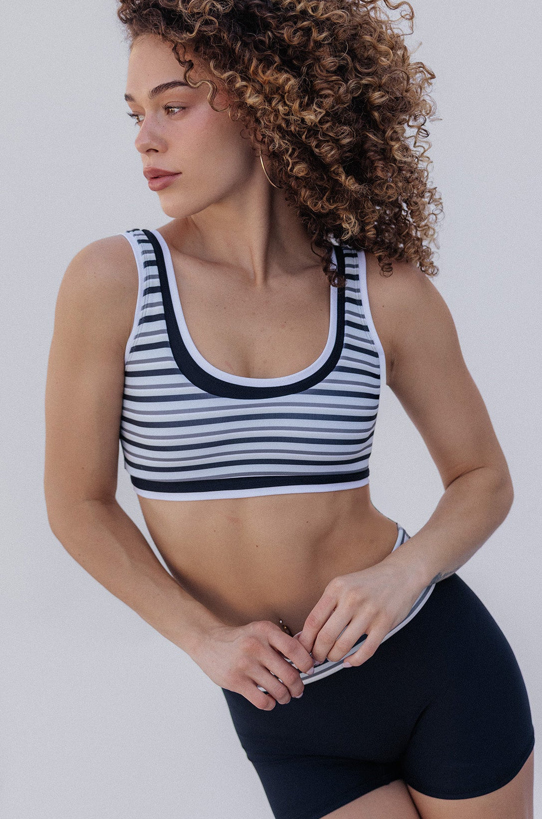 A woman with curly brown hair wearing a black and white striped sports bra and black athletic shorts, posing outdoors against a white wall.
