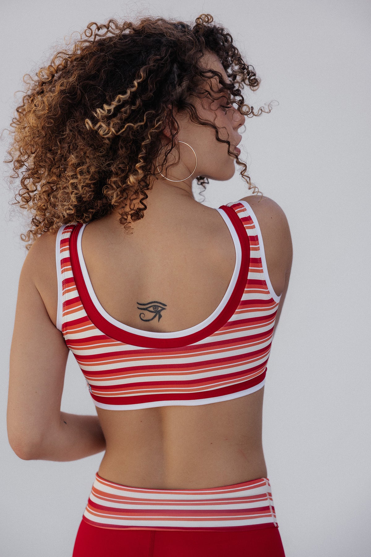 women, A woman with curly brown and blonde hair is shown from behind wearing a red and white striped sports bra and matching red bottoms against a light gray background.