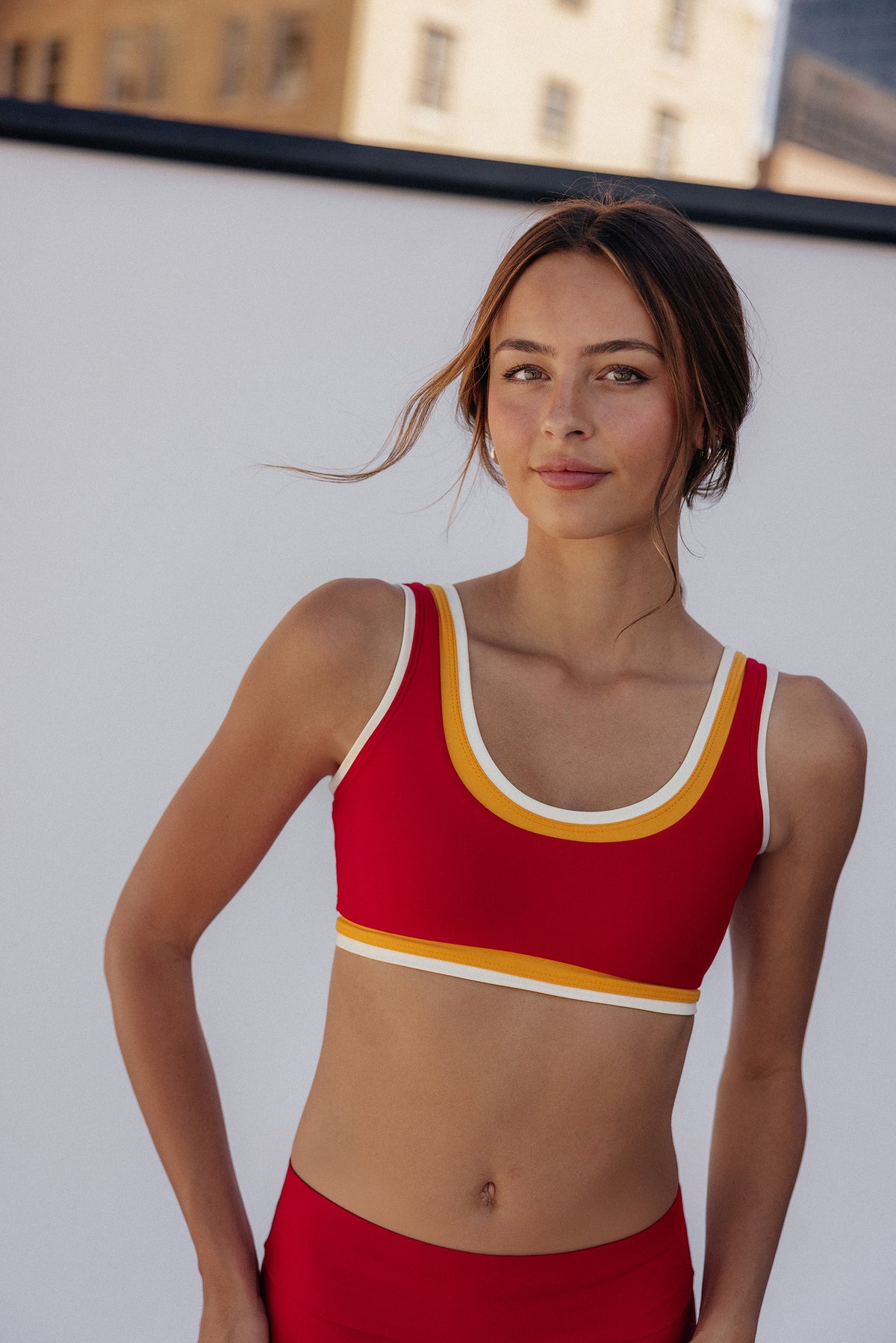 women, A woman modeling a red sports bra with yellow and white trim, paired with matching red bottoms, set against a bright white wall with buildings in the background.