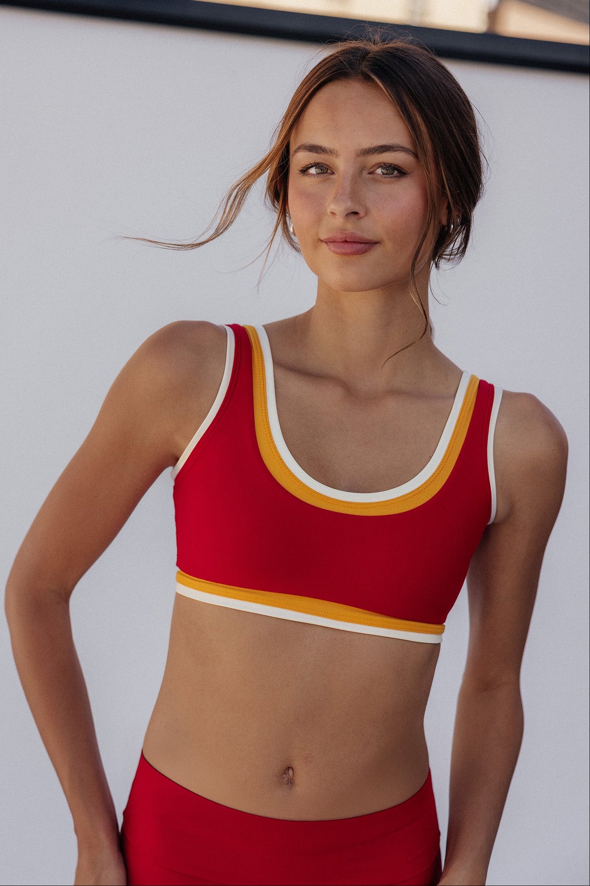 A woman modeling a red sports bra with yellow and white trim, paired with matching red bottoms, set against a bright white wall with buildings in the background.