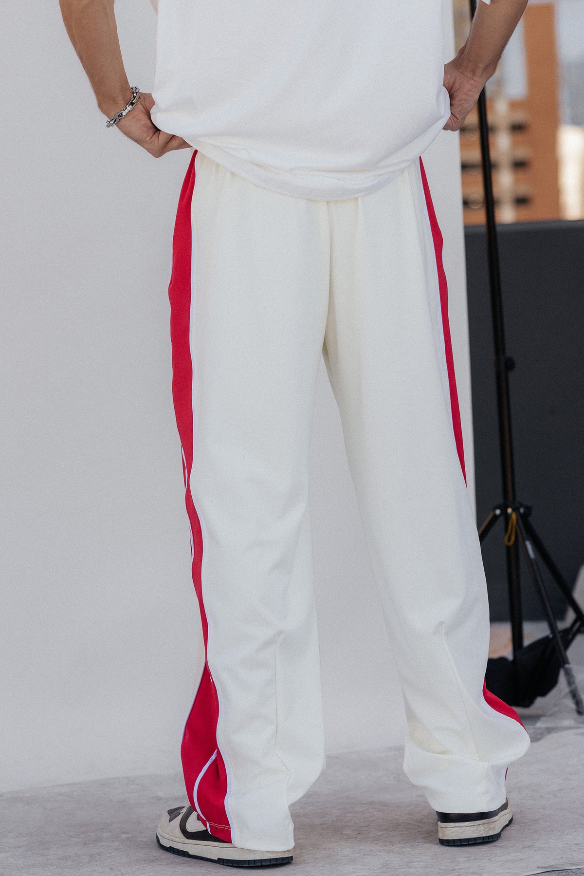 men, White wide-leg track pants with bold red side stripes, worn by a model from the back, paired with a white t-shirt and white sneakers