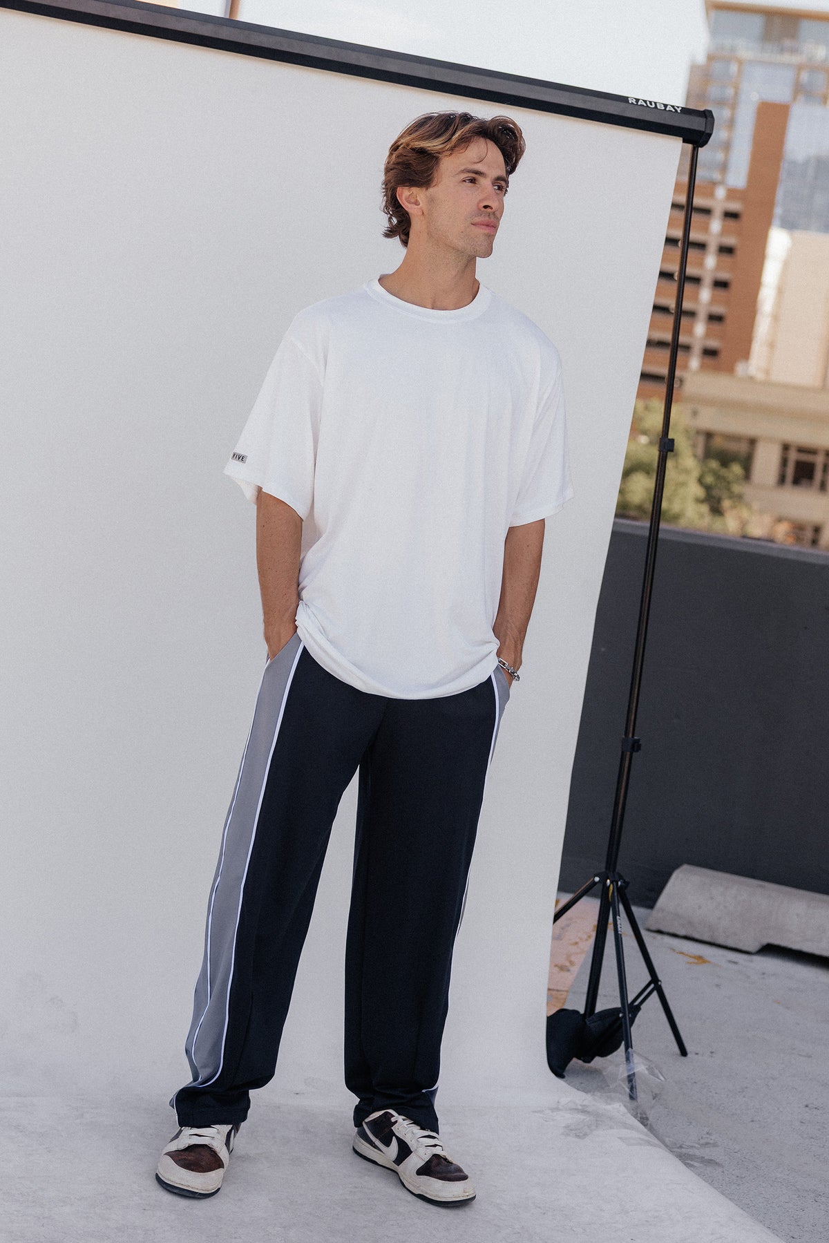 men, A man wearing an oversized white t-shirt and dark navy track pants with a light gray side stripe, standing in front of a white studio backdrop with a city skyline in the background.