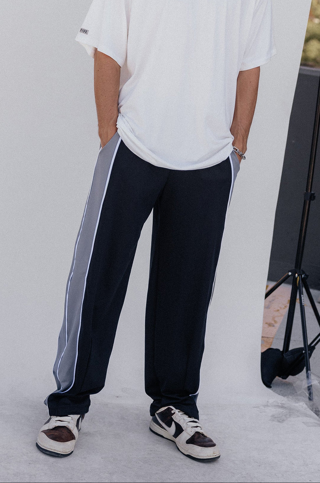 men, A man wearing an oversized white t-shirt and dark navy track pants with a light gray side stripe, standing in front of a white studio backdrop with a city skyline in the background.