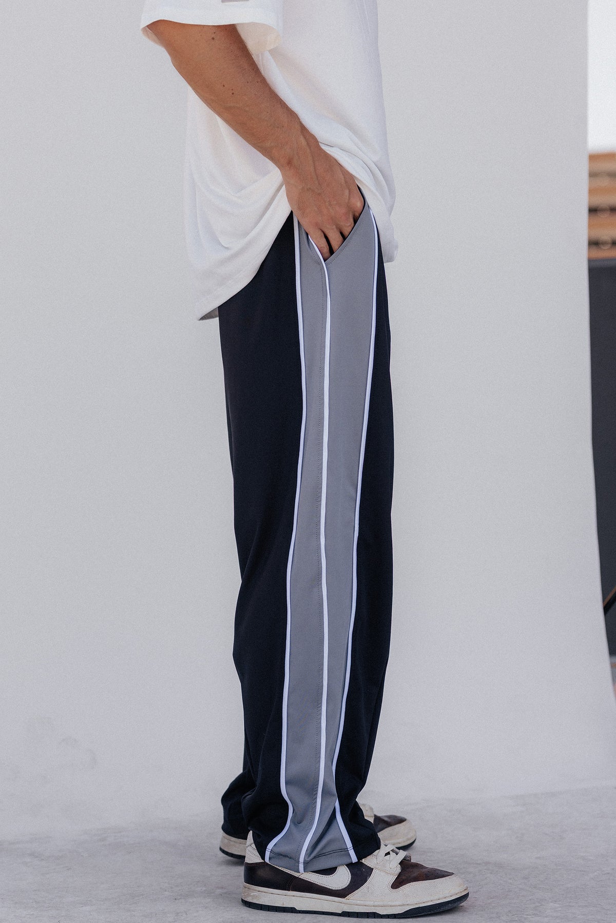 men, A person wearing loose dark navy track pants with a wide light gray side stripe and white piping, paired with a white T-shirt and white-and-black sneakers, shown from the waist down against a light studio background.