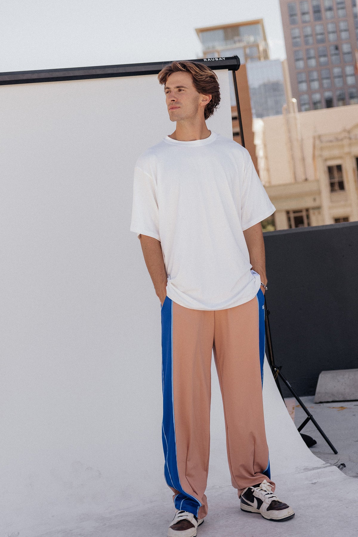 men, A man modeling a white oversized t-shirt and nude-colored track pants with blue side stripes, standing in front of a white studio backdrop with a city skyline in the background.