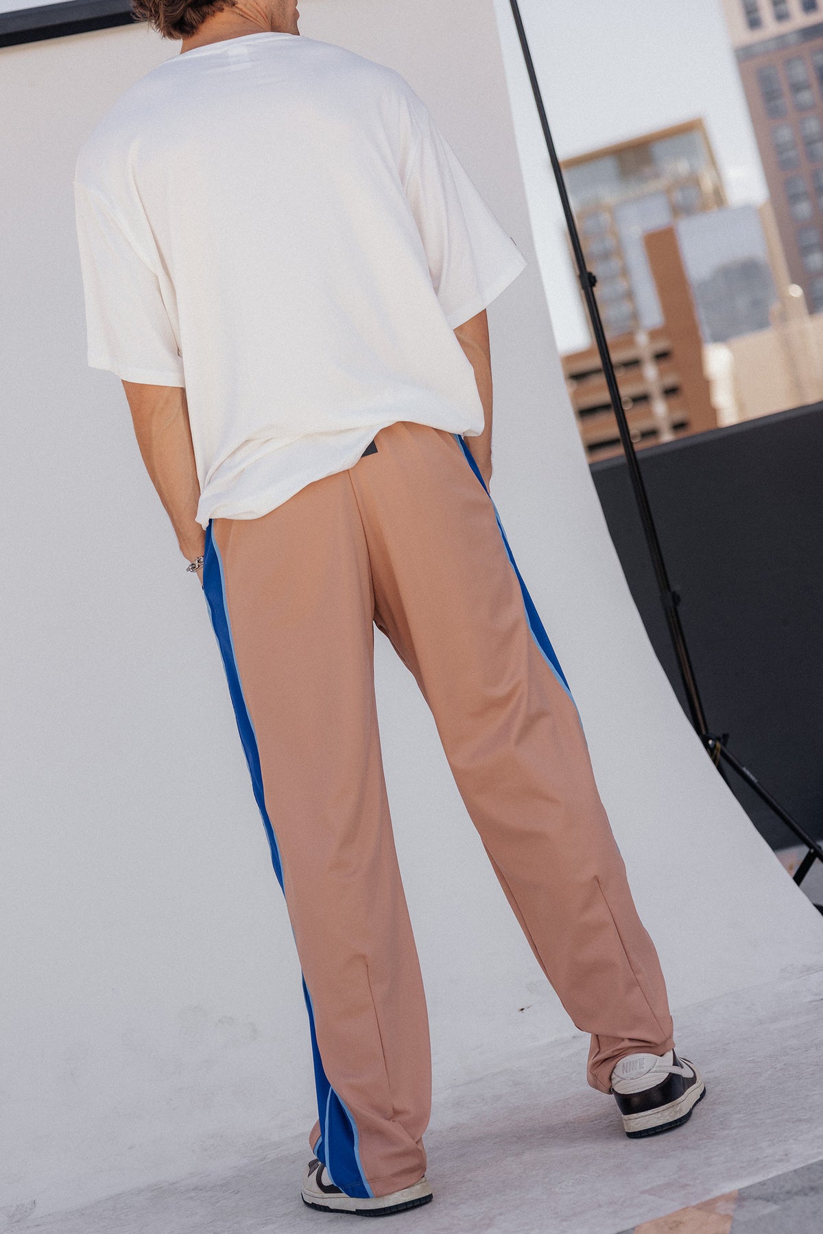 men, A person photographed from behind wearing loose-fit nude pants with bold blue side panels, a white oversized T-shirt, and black-and-white sneakers in an outdoor urban setting.