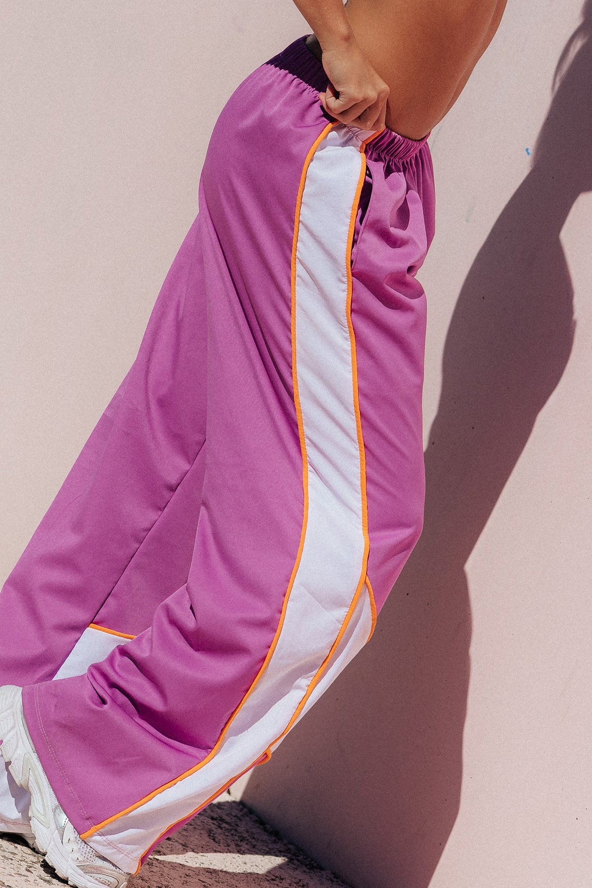 teen, A person wearing wide-leg athletic pants in a bright pink-purple color with a white side stripe bordered by orange piping, standing against a pale pink wall with a strong shadow.