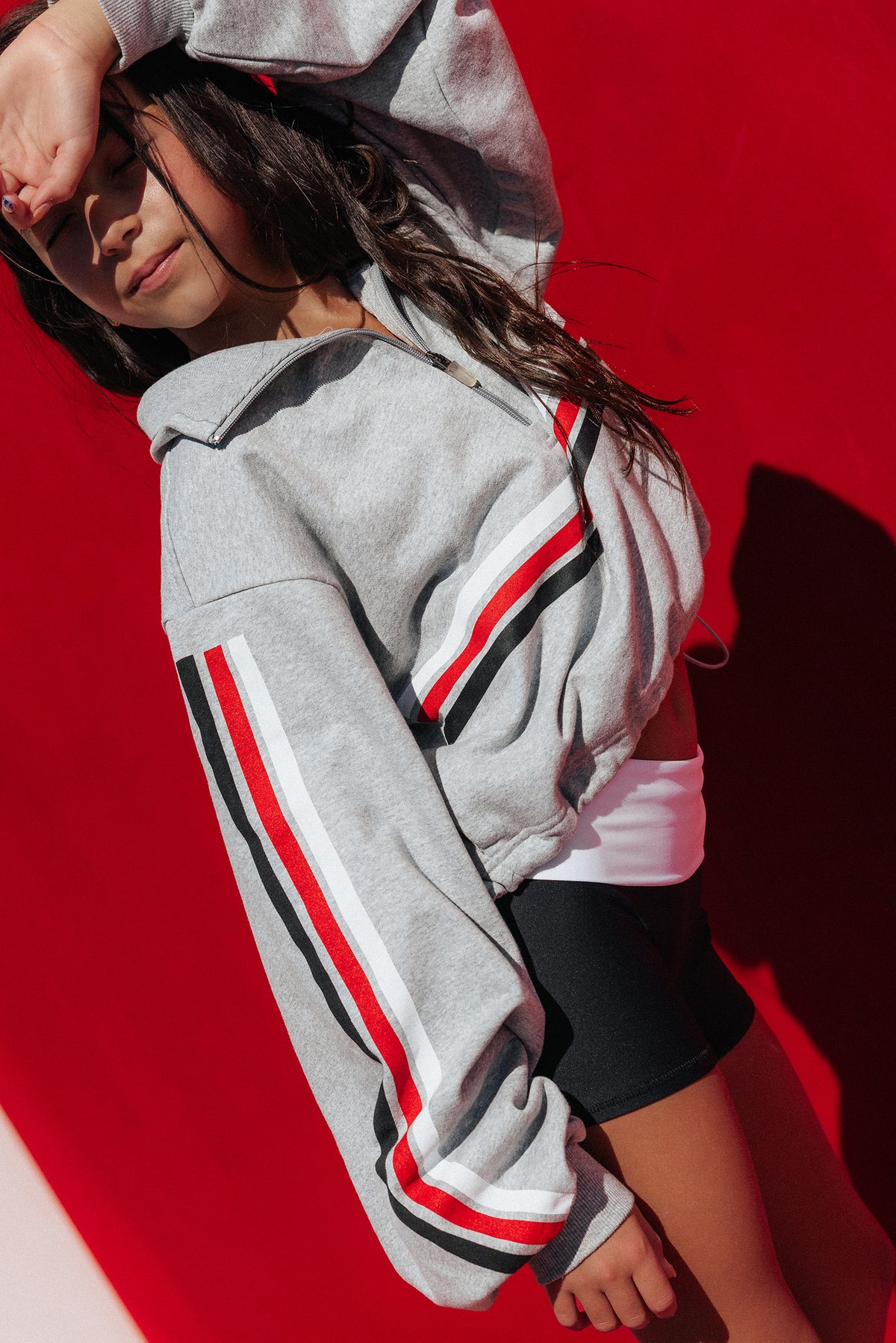 kids, A person wearing a cropped light gray hoodie with bold red, white, and black striped details on the sleeves and chest, paired with black shorts, posed against a vivid red background.