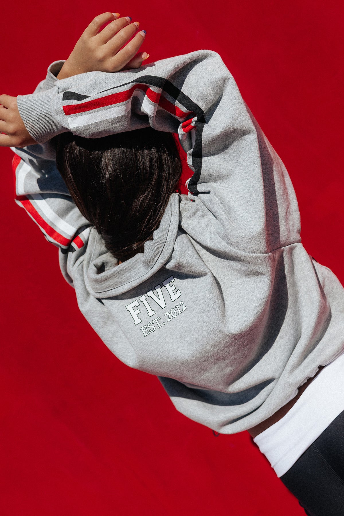 kids, A person wearing a light gray hoodie with red, white, and black striped sleeve details and 'FIVE EST. 2014' printed on the back, posed against a solid red background.