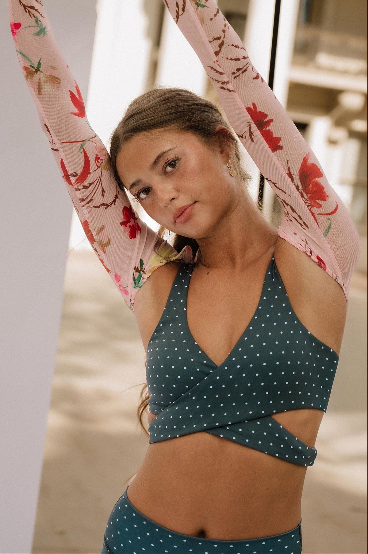 teen, in a grey blue and white polka dot activewear set stretching with her arms raised, wearing a matching floral long-sleeve top against a bright, sunlit outdoor background.