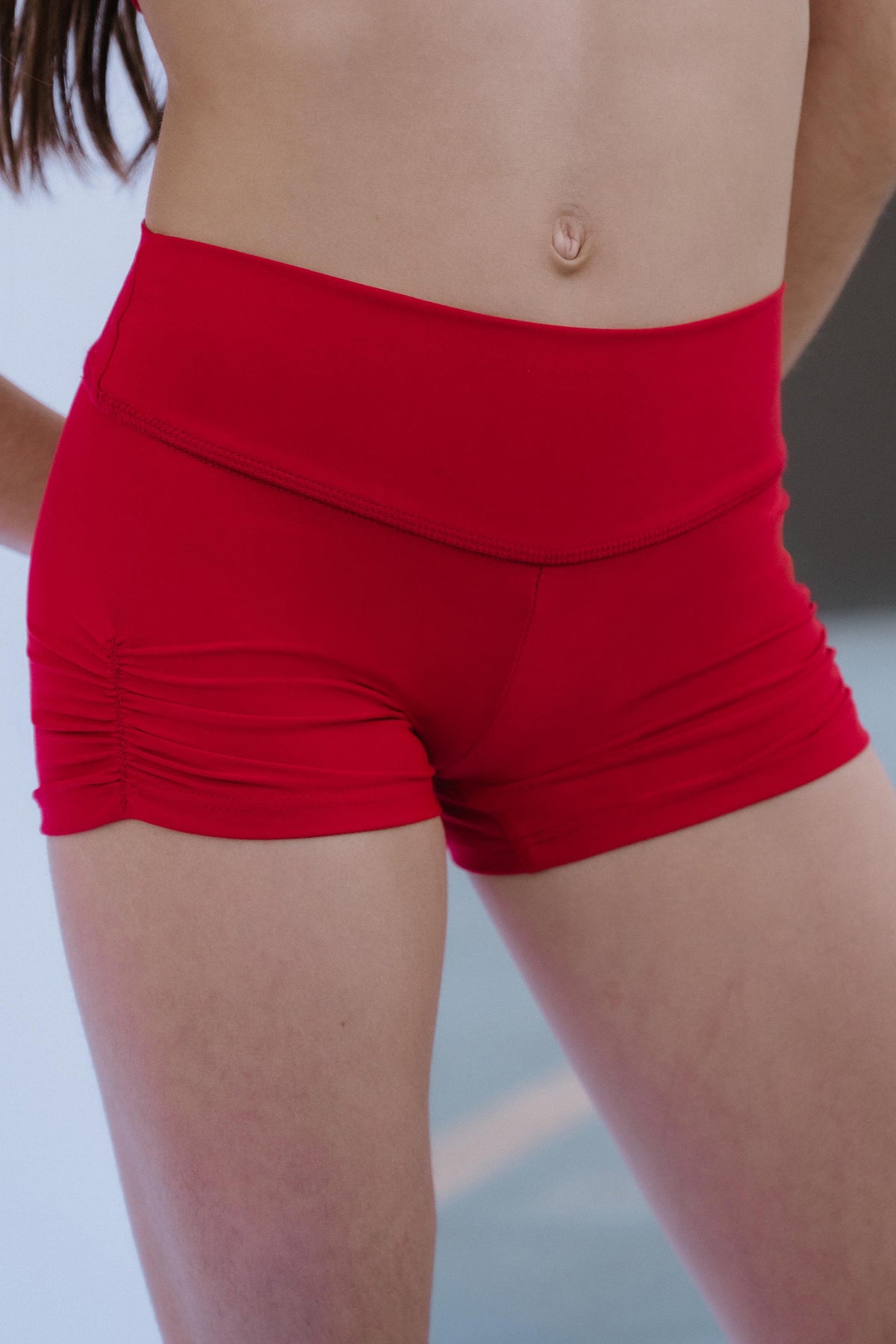 kids, A close-up of a model wearing bright red high-waisted athletic shorts with a gathered ruched detail at the front hem, shown against an outdoor blurred background.