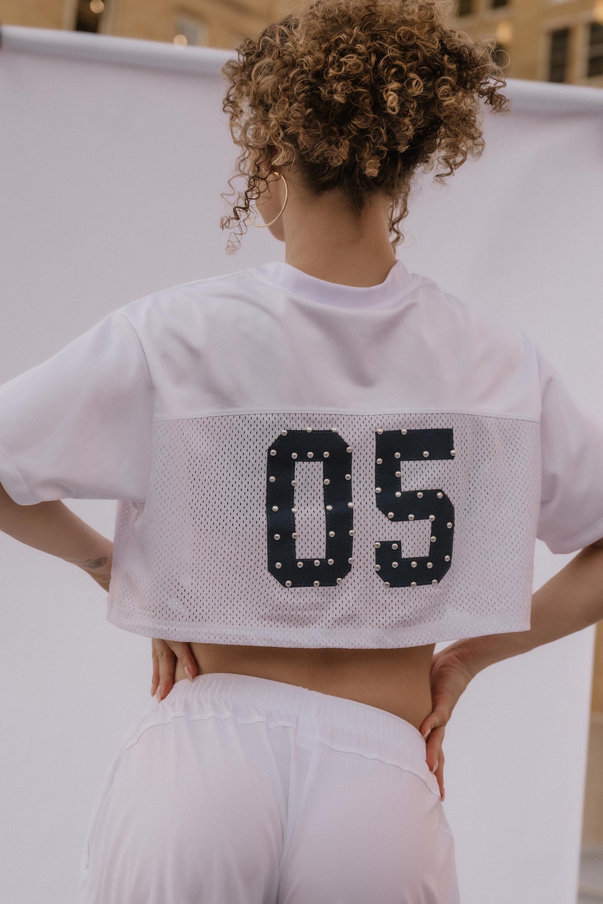 A person shown from behind wearing a white cropped athletic jersey with a large black '05' printed on the back, paired with white shorts, against a white backdrop.