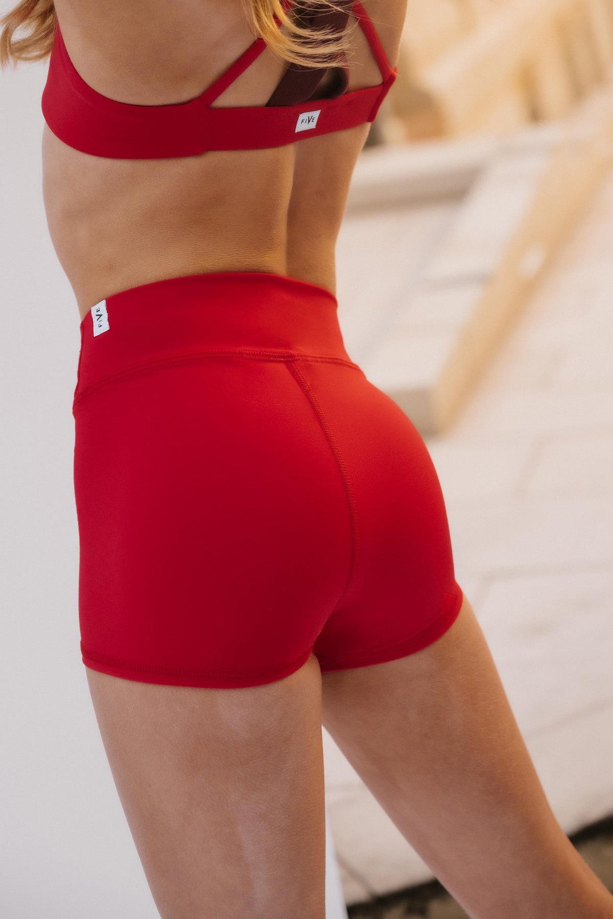 kids, A close-up back view of a model wearing bright red athletic shorts and a matching red sports bra, with a small white brand tag visible on the waistband.