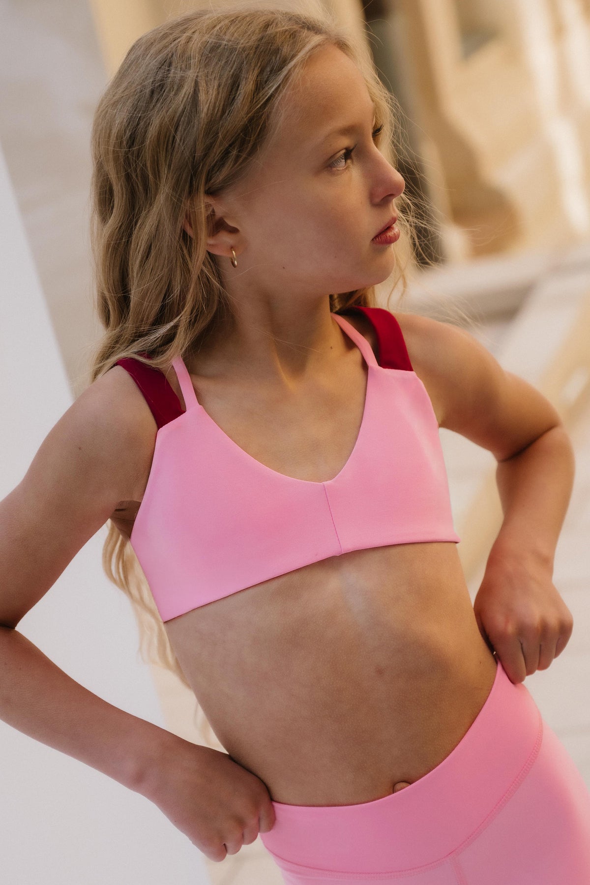 kids, A young girl wearing a light pink sports bra with contrasting dark red shoulder straps and matching pink leggings, posing in a brightly lit indoor setting.