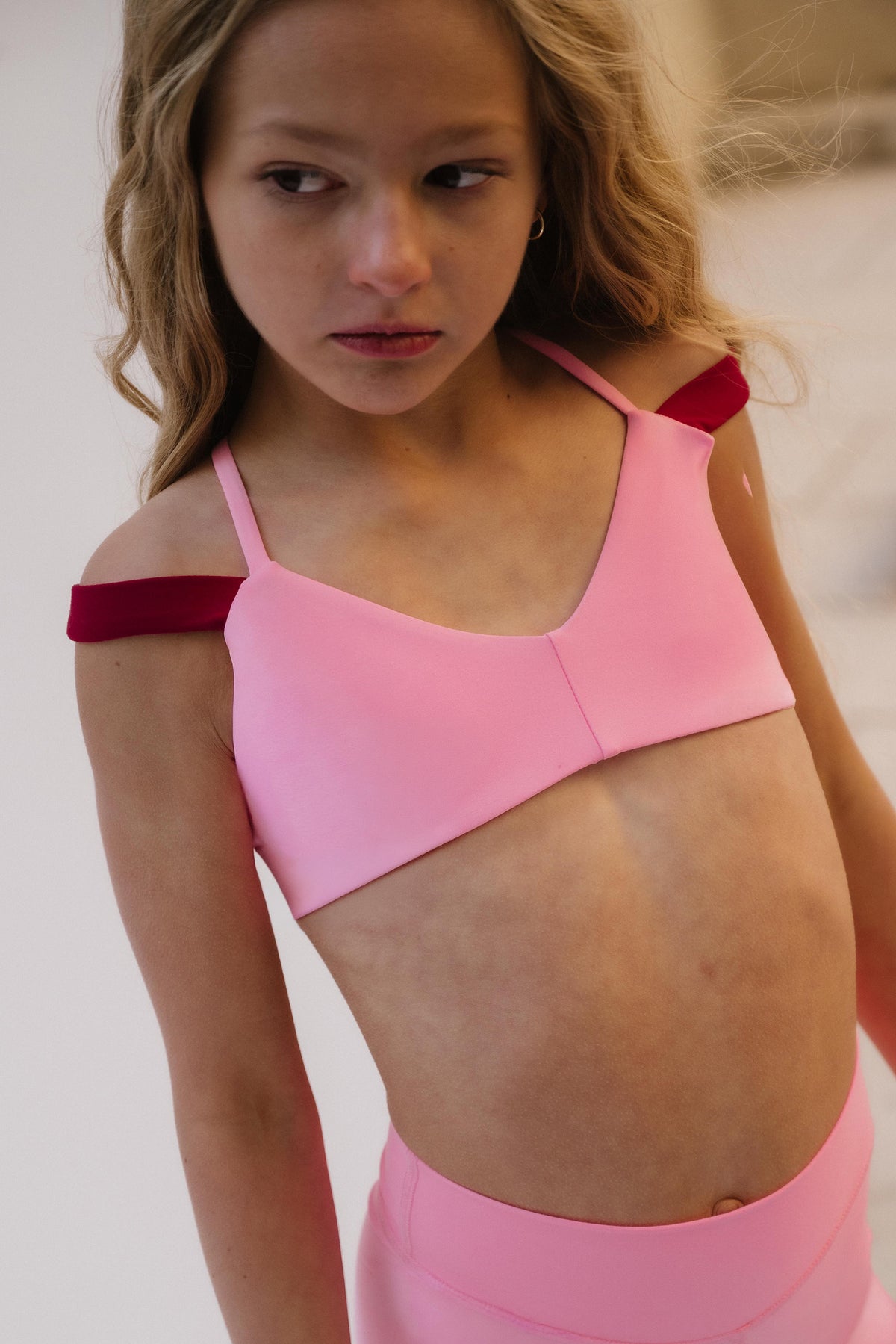 kids, A young girl wearing a pink sports bra with contrasting dark red shoulder straps and matching pink bottoms, photographed in a softly lit indoor setting.