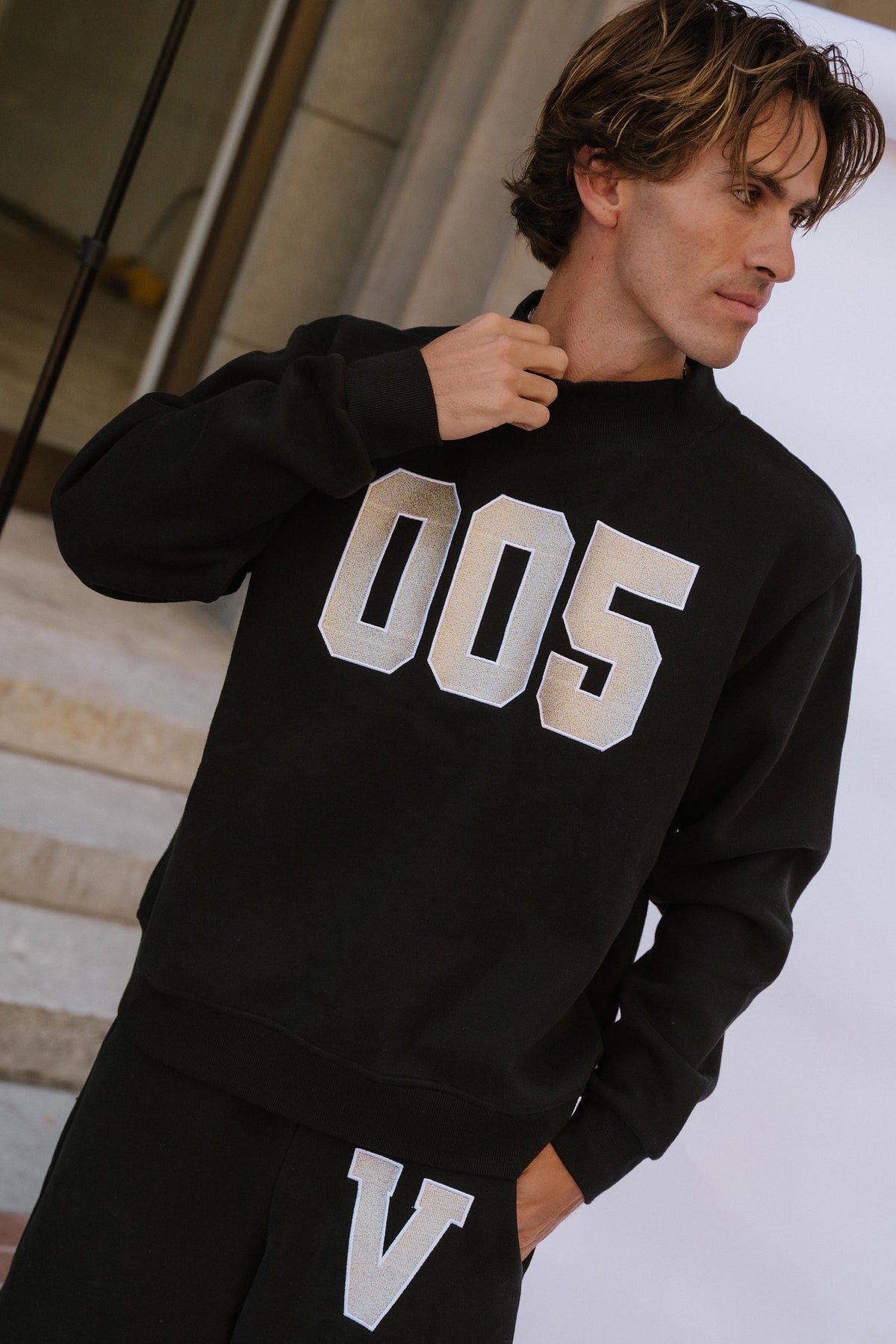 mens, A man wearing a black sweatshirt with large white '005' lettering on the front and matching black pants with a white 'V' graphic, photographed in an indoor setting with steps in the background.