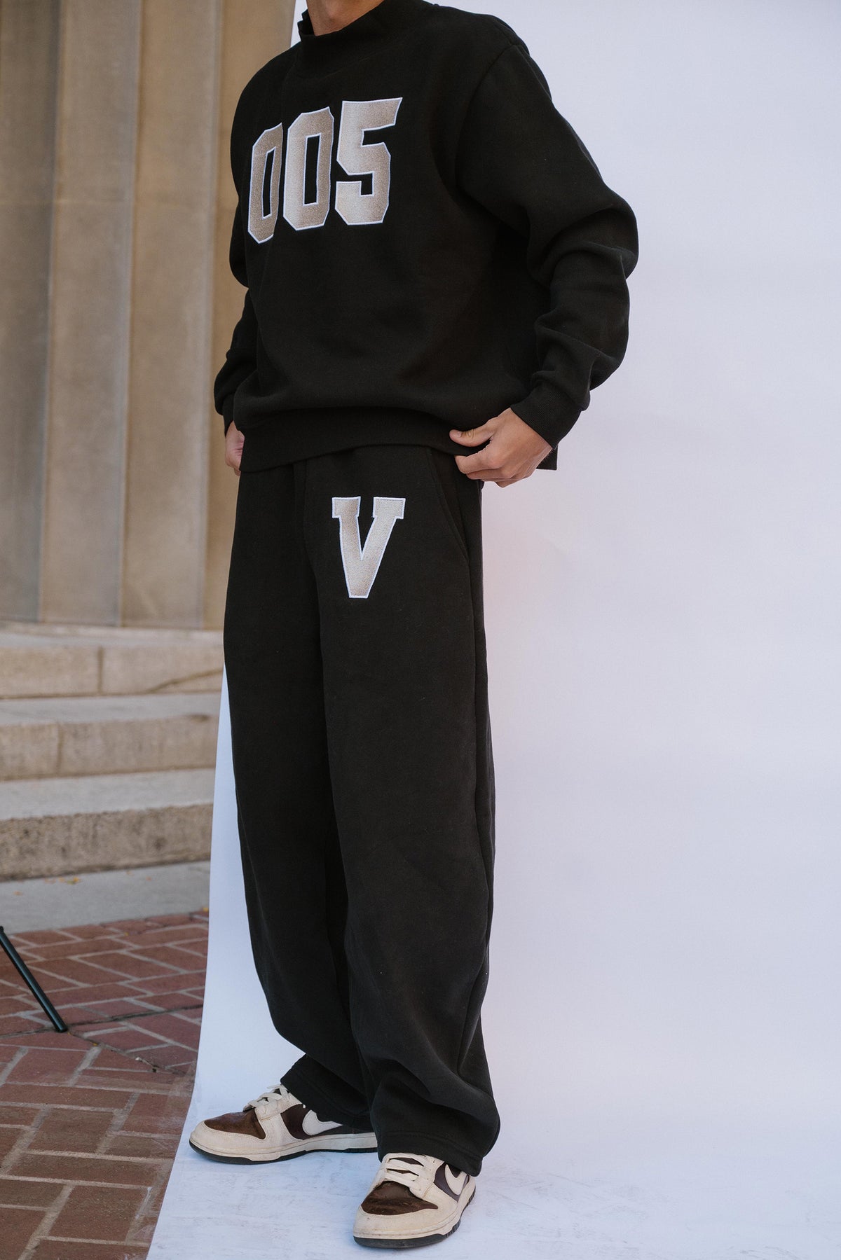 mens, A person wearing a black oversized sweatshirt with large white '005' lettering on the chest and matching black sweatpants with a large white 'V' on the thigh, standing against a white studio backdrop with stone steps visible on the left.