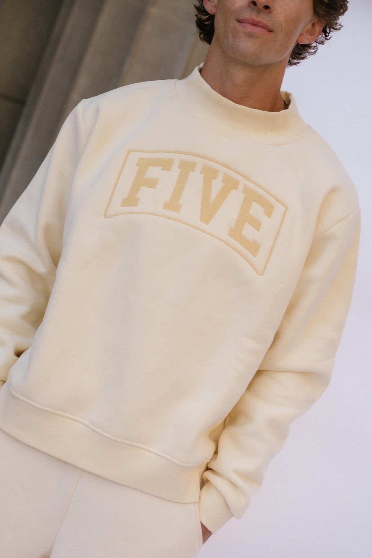 men, A close-up of a man wearing a cream-colored crewneck sweatshirt with 'FIVE' embroidered in a matching beige tone on the chest.