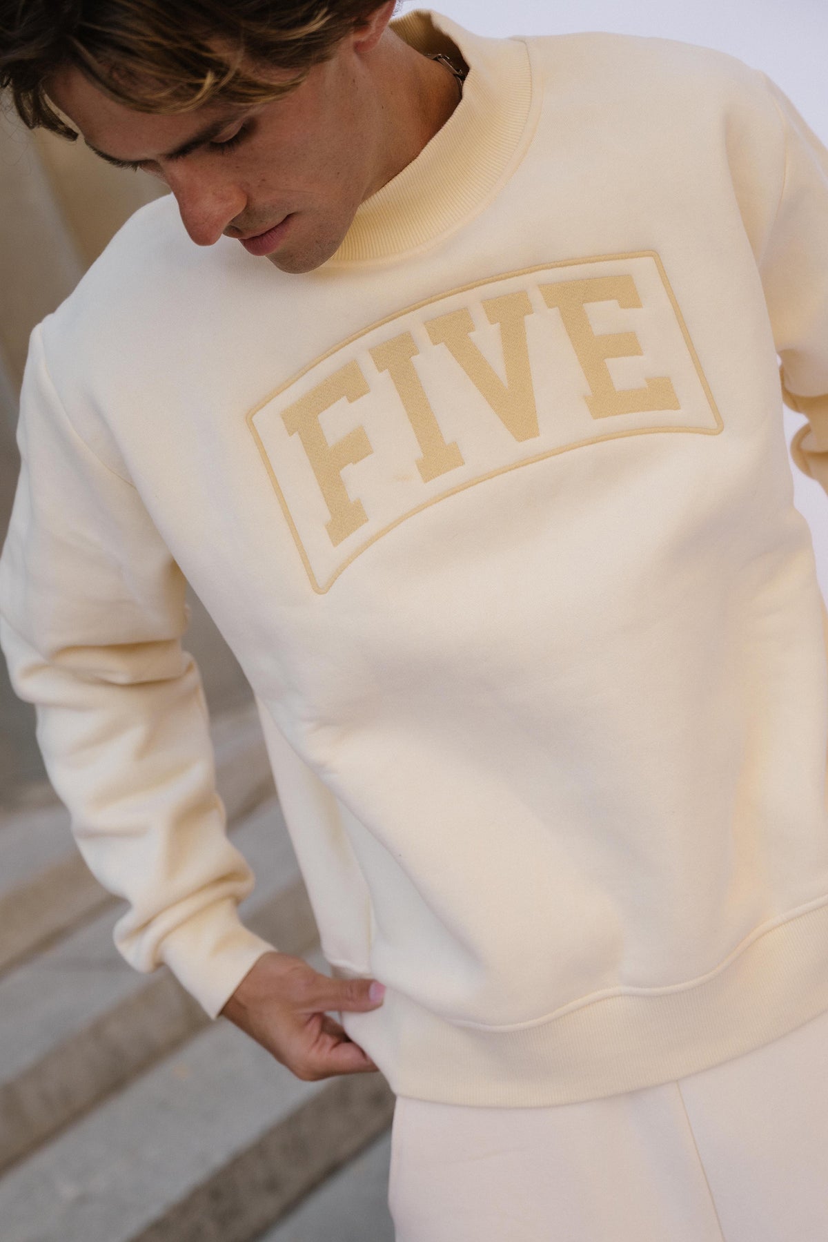 men, A man wearing a cream-colored crewneck sweatshirt with a tonal beige 'FIVE' graphic on the chest, standing on stone steps.