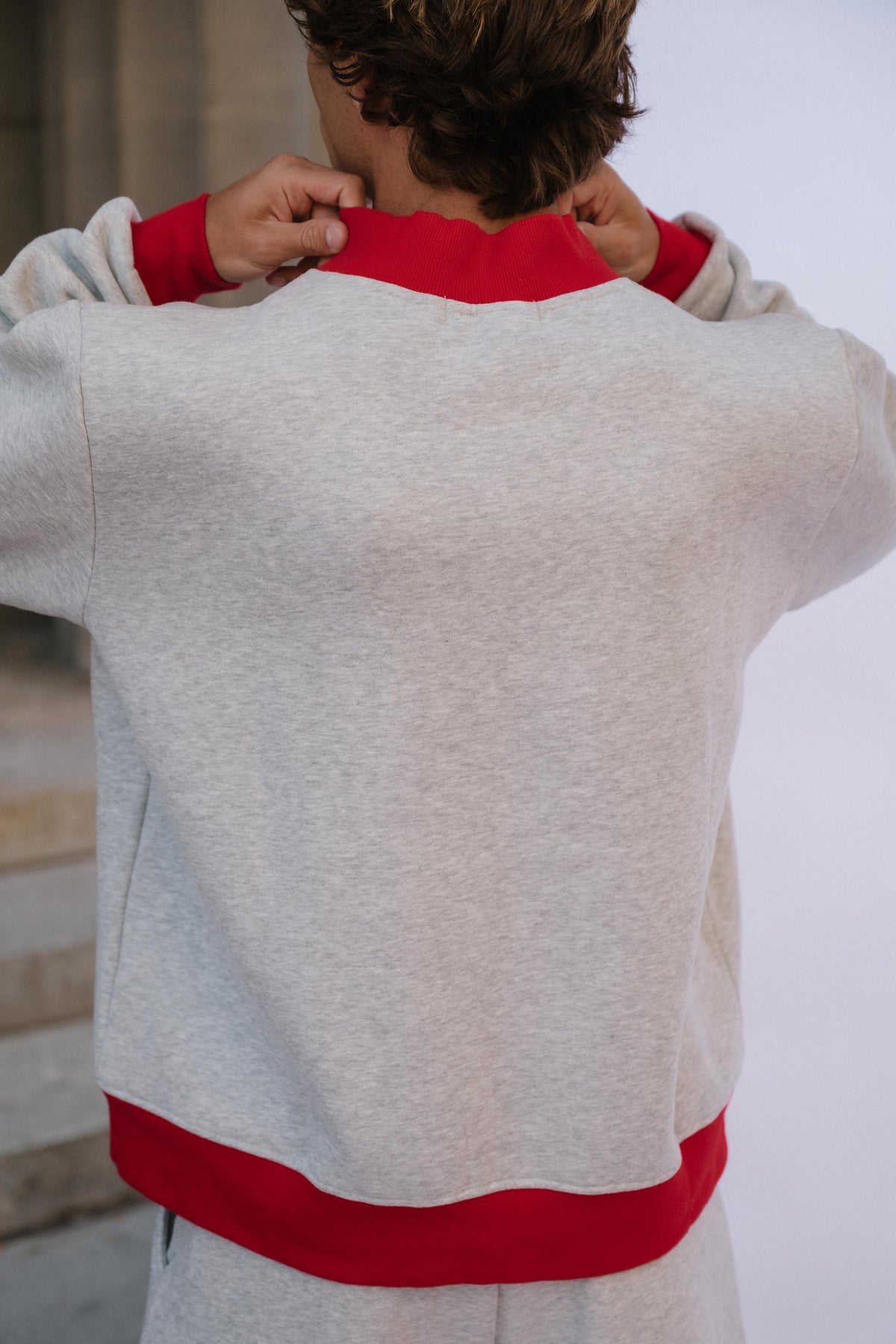 mens, A person viewed from behind wearing a light heather gray sweatshirt with contrasting red ribbed collar, cuffs, and hem, with matching gray pants partially visible.