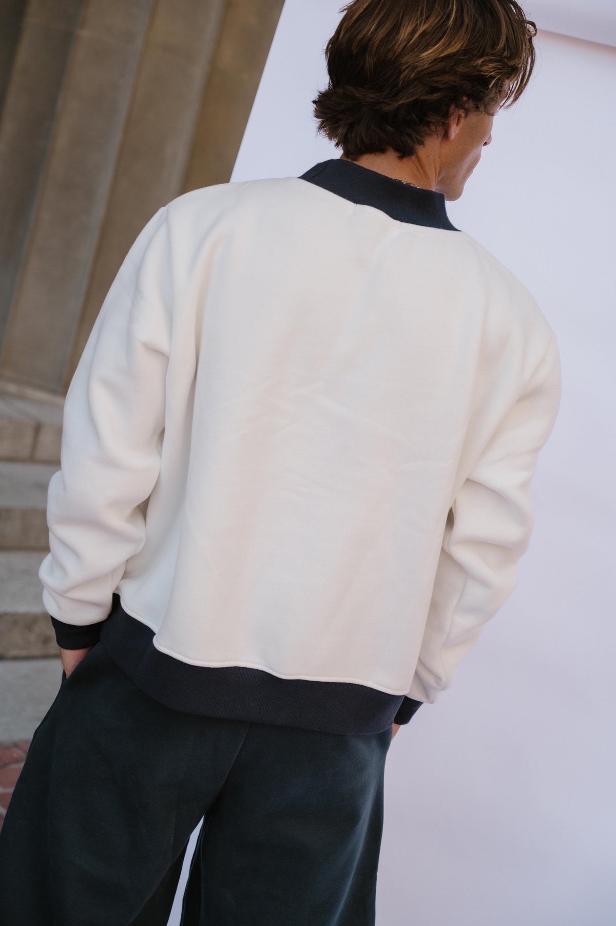 mens, A person shown from the back wearing a cream-colored bomber-style sweatshirt with dark navy ribbed collar, cuffs, and hem, paired with dark pants.