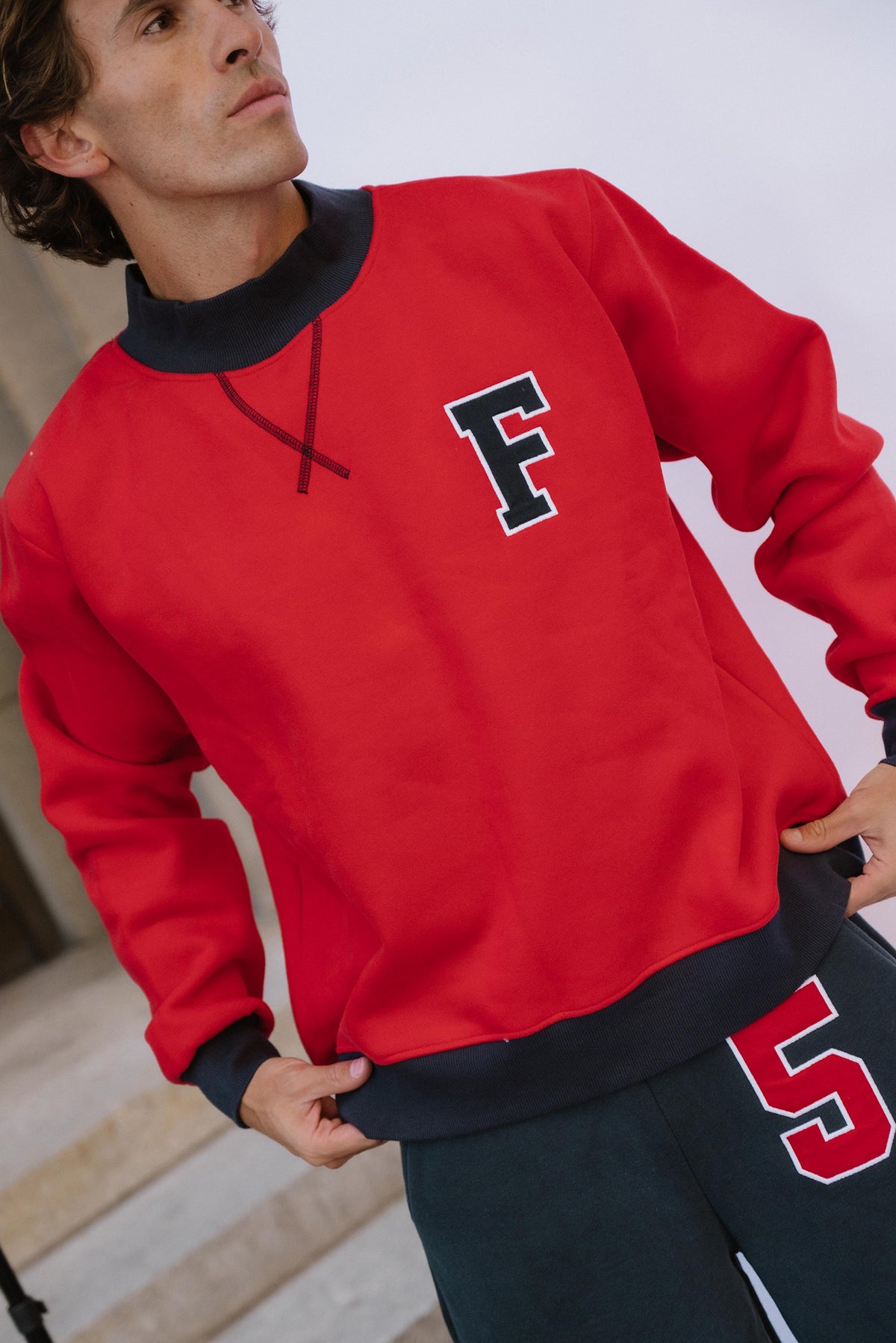 mens, A man wearing a bright red crewneck sweatshirt with a navy blue collar, cuffs, and hem, featuring a navy blue block letter 'F' with a white outline on the left chest. He is also wearing dark navy sweatpants with a red and white '5' on the thigh.
