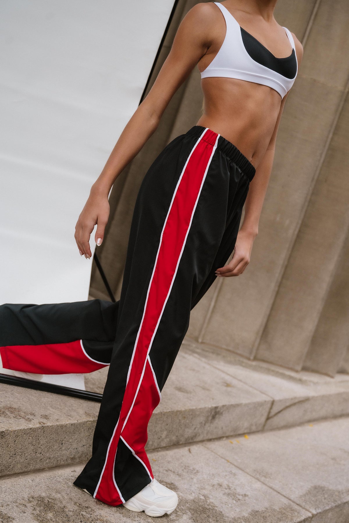kids, A model wearing black wide-leg athletic pants with a bold red and white side stripe, paired with a white and black sports bra, standing on concrete steps.