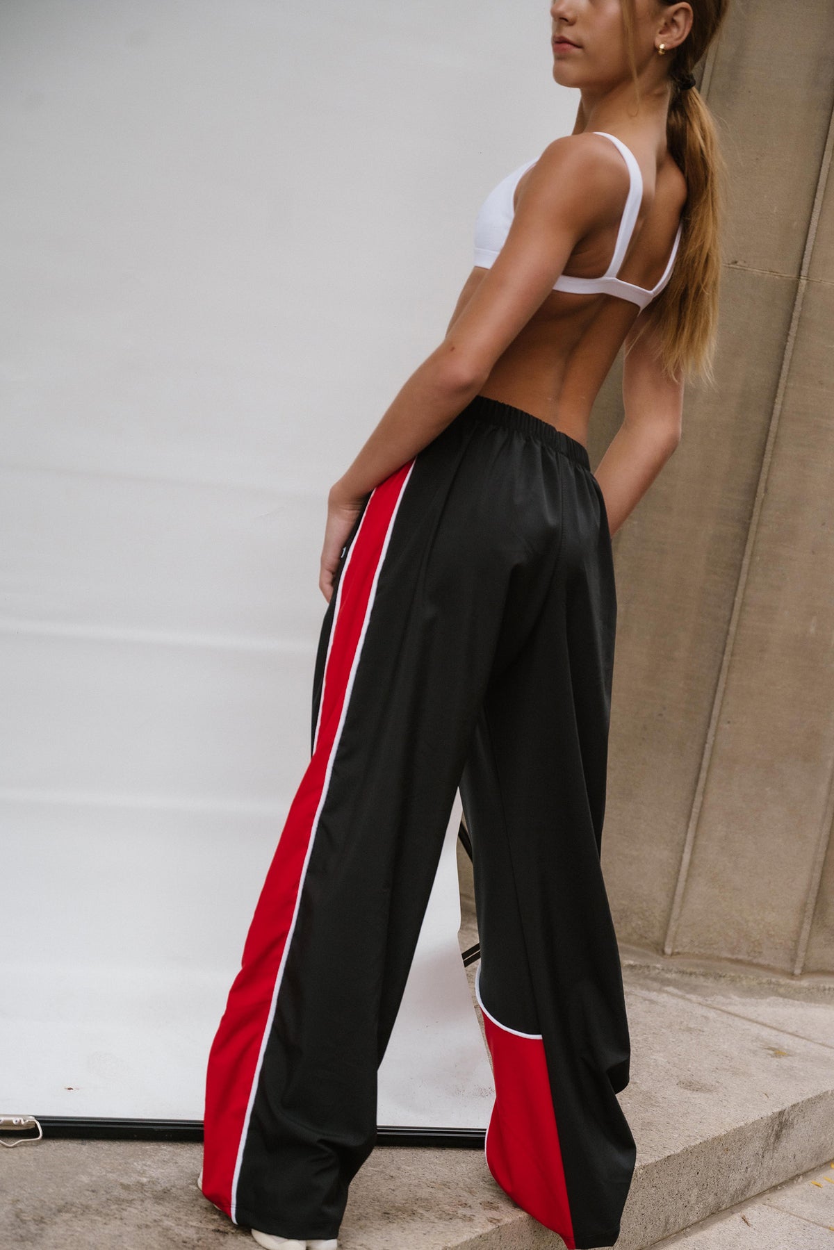 kids, kid wearing black wide-leg athletic pants with bold red and white side stripes, paired with a white sports bra, standing against a light gray wall.