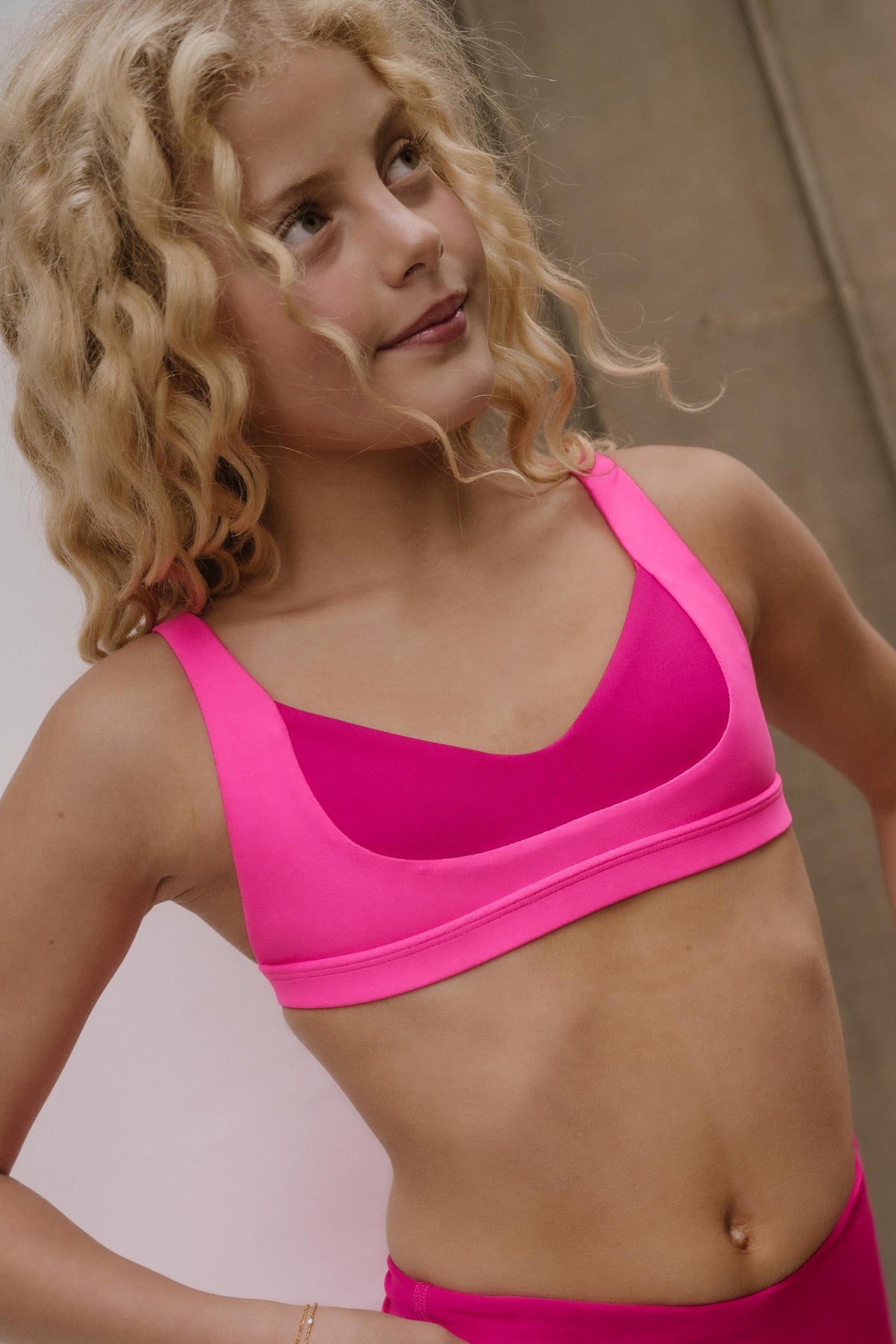 kids, A close-up shot of a young girl with curly blonde hair wearing a bright hot pink sports bra and matching pink bottoms, looking off-camera against a neutral background.