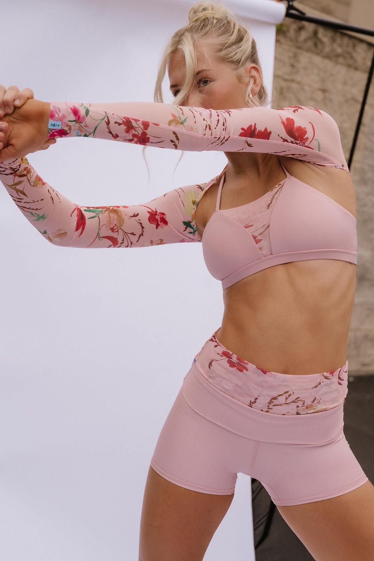 teen, modeling a coordinated activewear set consisting of a light pink sports bra, matching high-waisted shorts, and a long-sleeve floral print arm warmer, posed against a white studio backdrop.