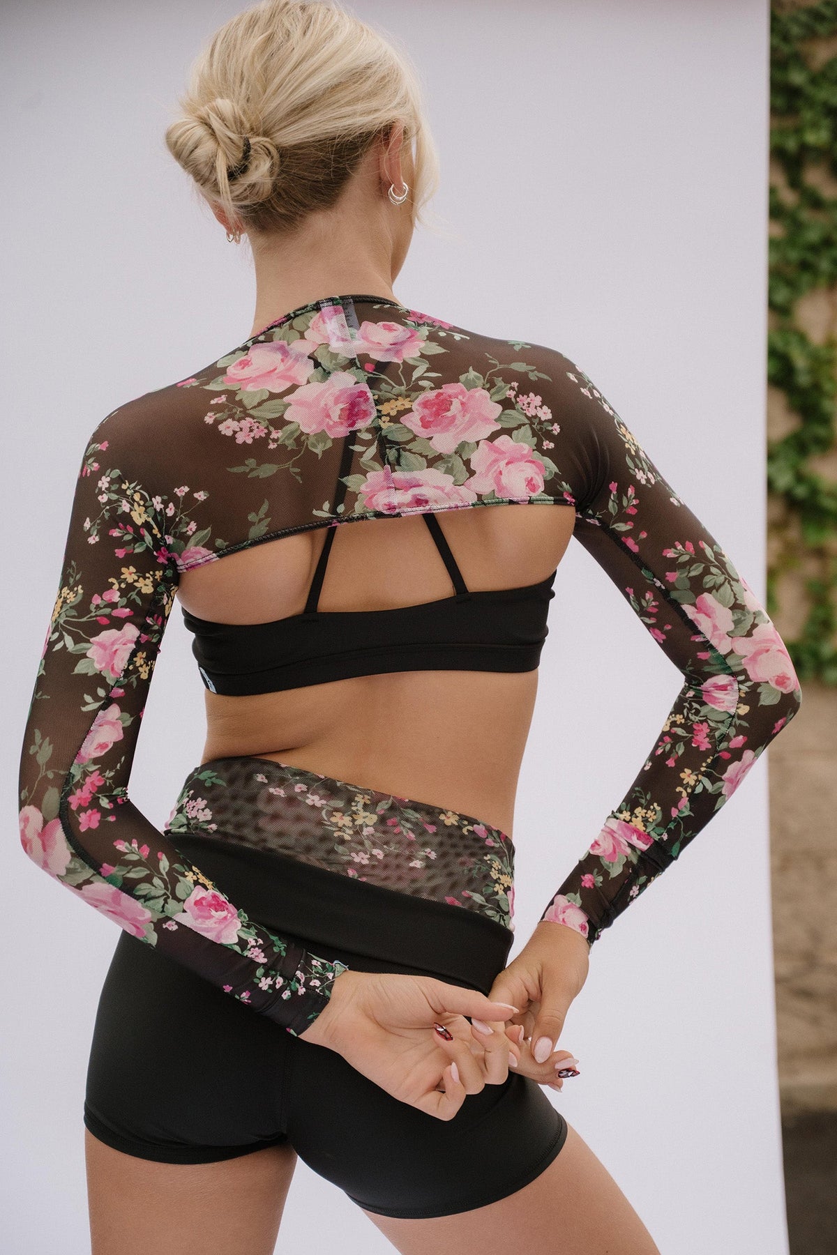 teen, shown from the back wearing a sheer long-sleeve floral athletic top with a black sports bra and black shorts, standing against a light studio backdrop.