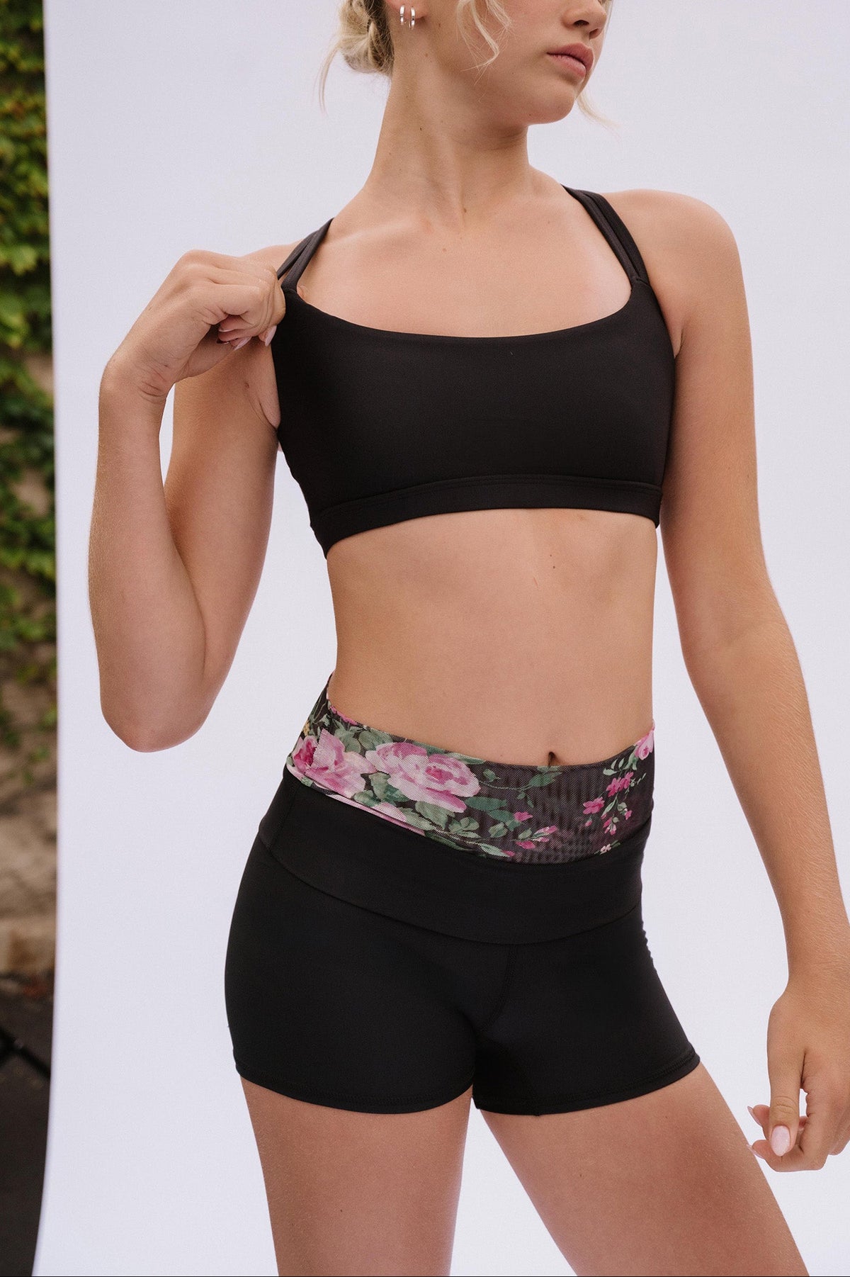 teen, blonde model wearing a black sports bra and matching black athletic shorts with a pink floral waistband, standing against a white studio backdrop.