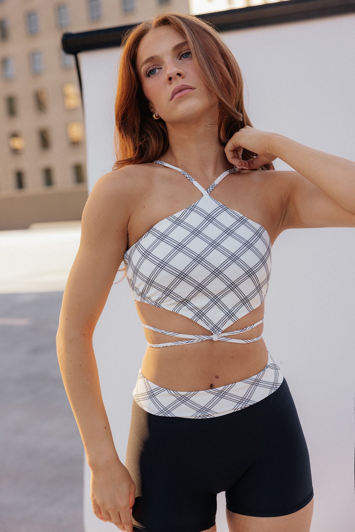 teen, girl modeling a white and gray plaid halter crop top with side cutouts and matching plaid waist ties, paired with black bike shorts, photographed outdoors against a white backdrop and building.