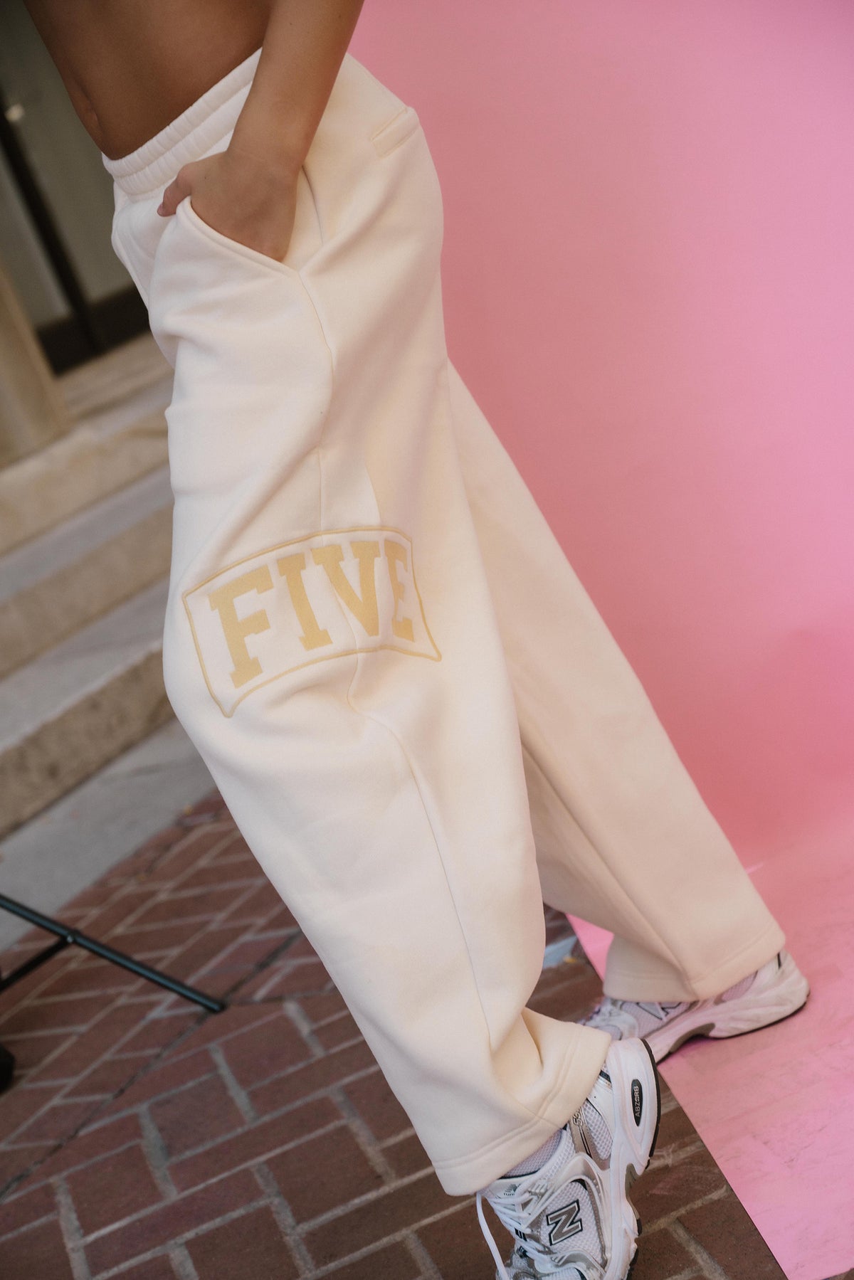 teen, A person wearing cream sweatpants with a gold 'FIVE' graphic on the leg, paired with white sneakers, standing against a pink backdrop.