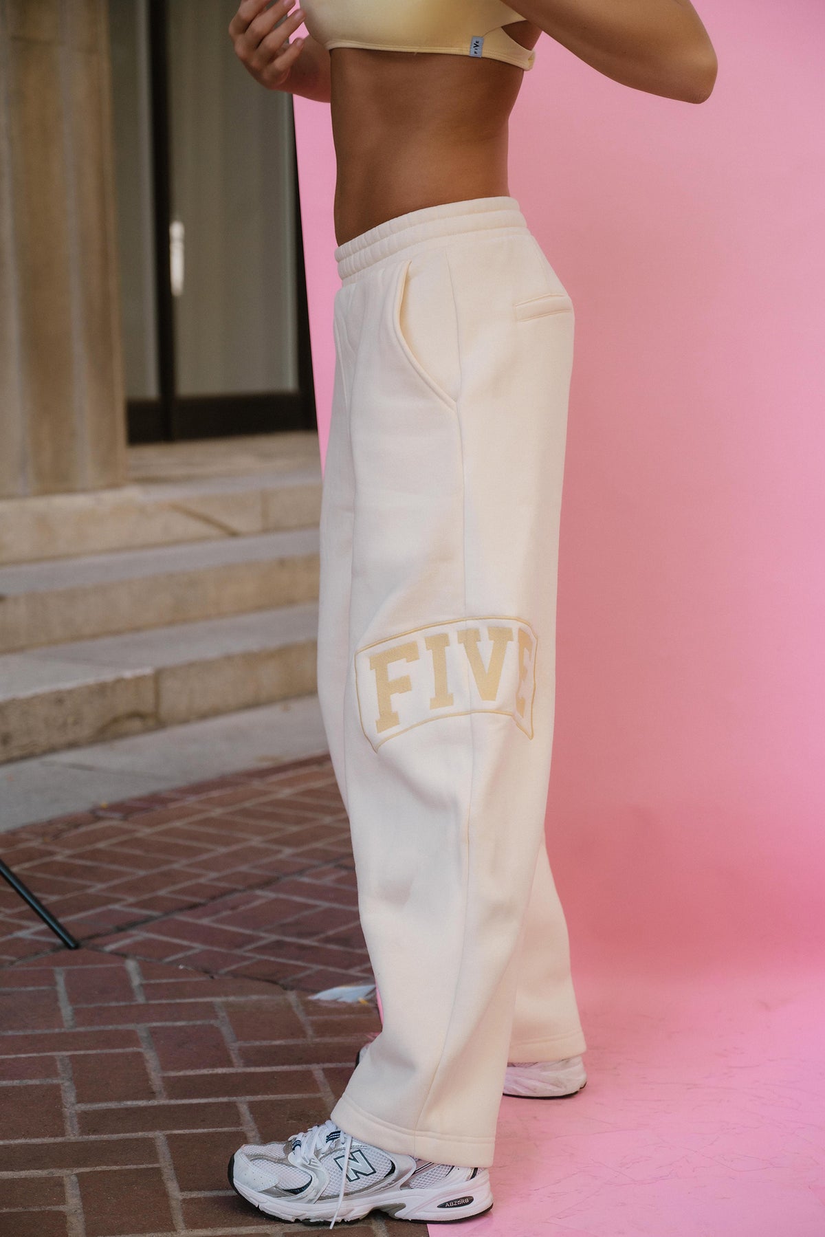 teen, A person wearing cream sweatpants with a gold 'FIVE' graphic on the leg, paired with a beige sports bra and white sneakers, standing against a pink backdrop.