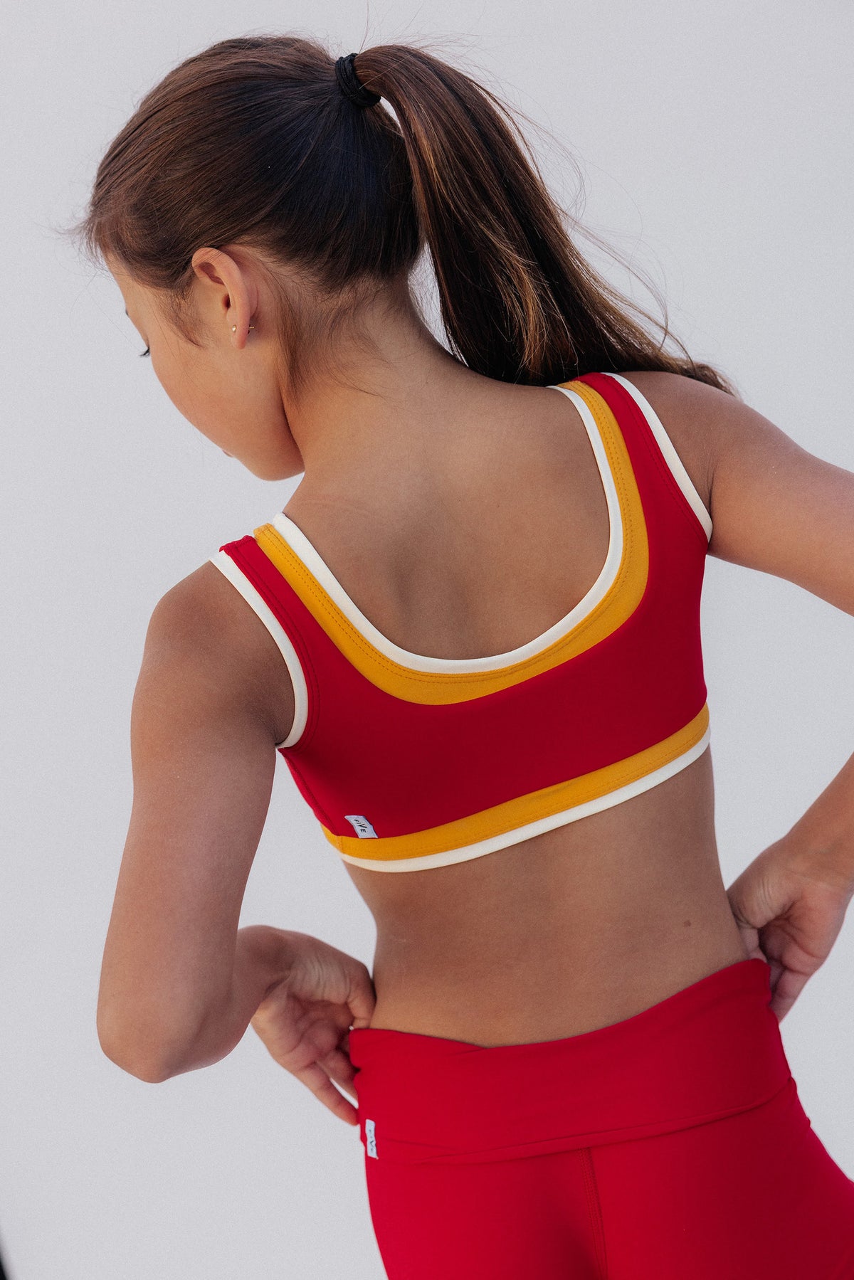 kids, A young girl modeling a red two-piece athletic set consisting of a scoop-neck crop top with yellow and white trim, and matching red athletic shorts, against a light gray studio background