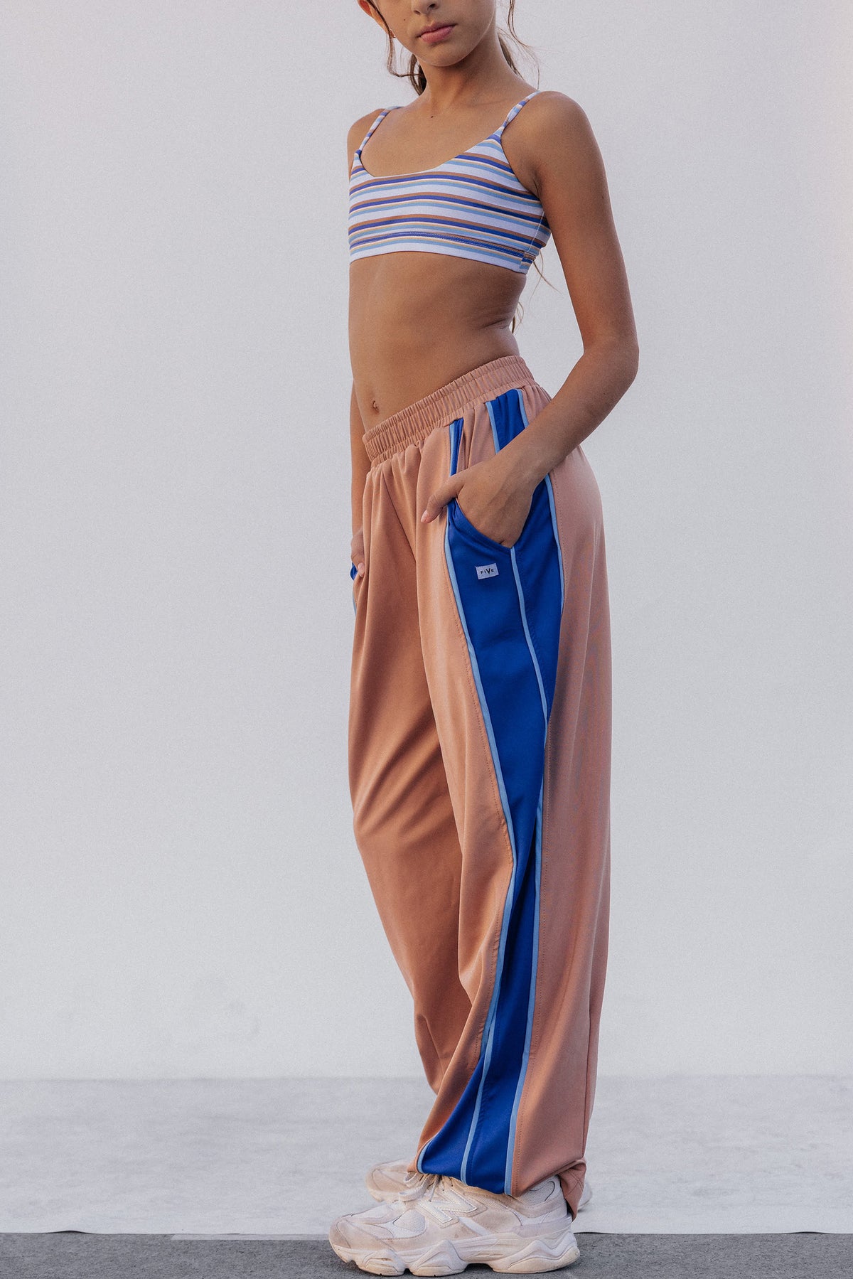 kids, A model wearing loose, wide-leg sweatpants in a dusty peach color with a bold blue side stripe, paired with a striped crop top and white sneakers, set against a plain light gray studio background.