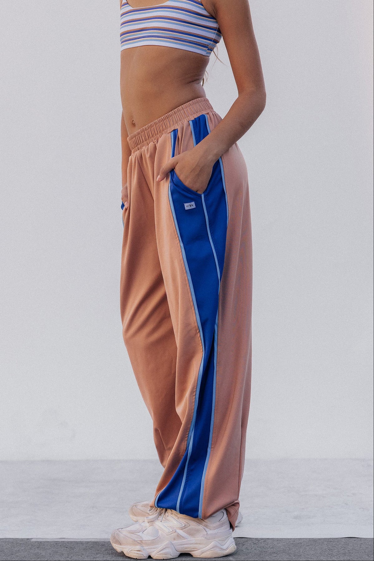 kids, A model wearing loose, wide-leg sweatpants in a dusty peach color with a bold blue side stripe, paired with a striped crop top and white sneakers, set against a plain light gray studio background.