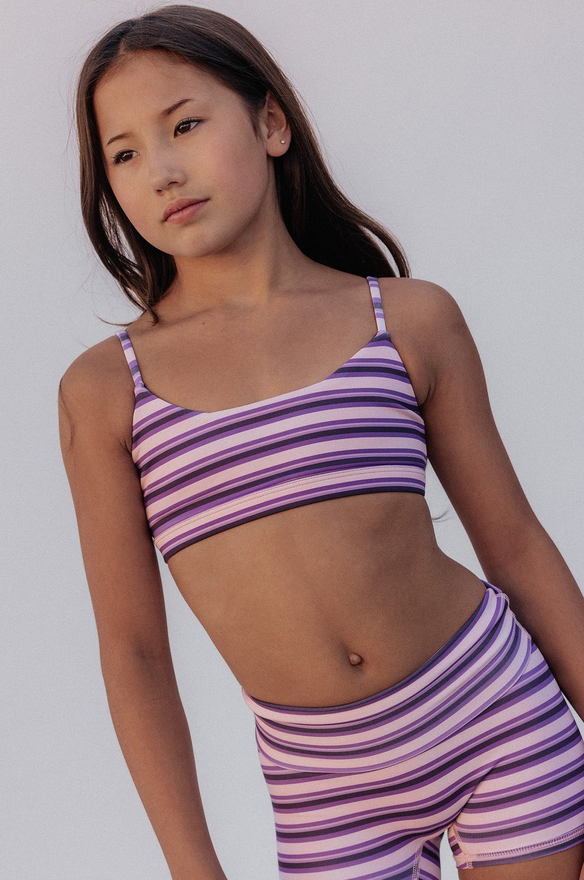 kids, A young girl modeling a matching two-piece activewear set with purple and white horizontal stripes, consisting of a spaghetti-strap crop top and high-waisted shorts, set against a plain light gray background.