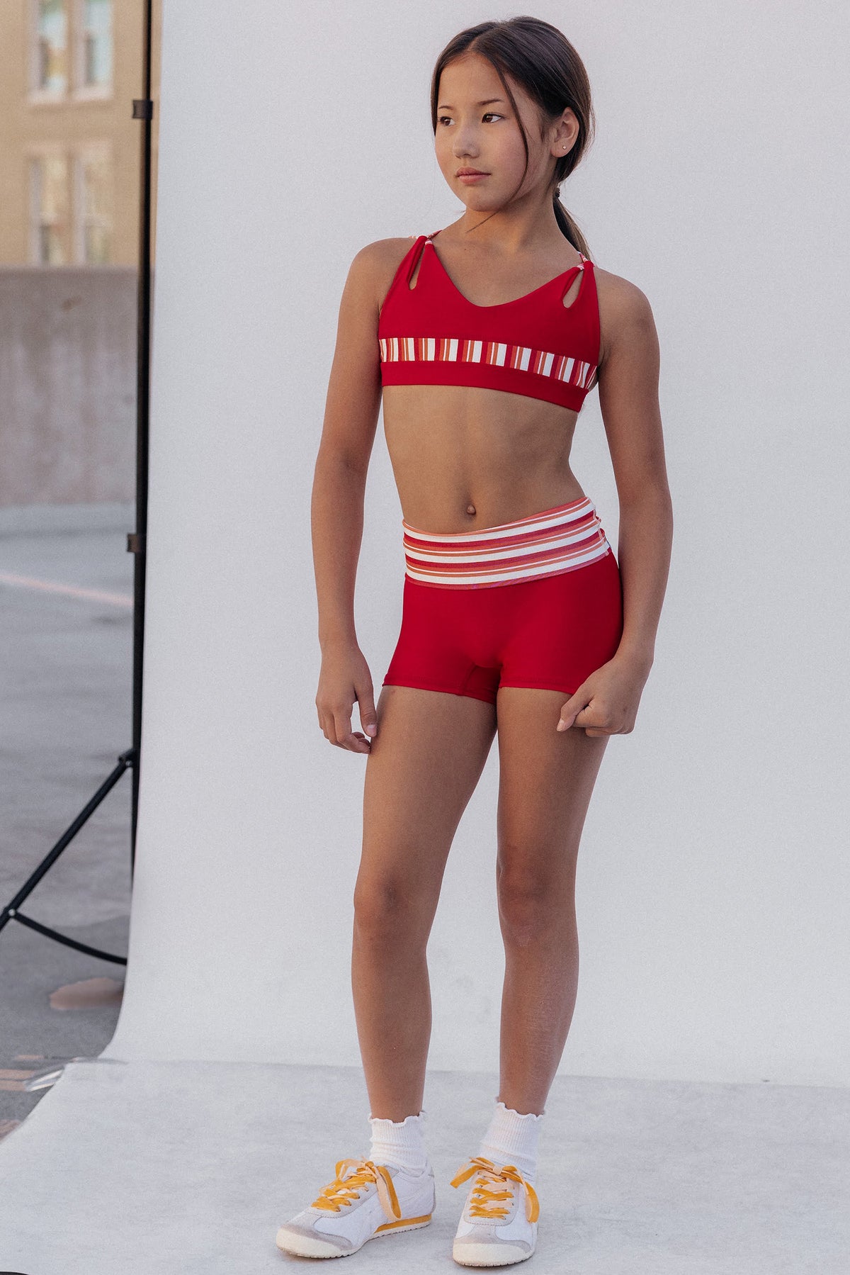 kids, A young girl modeling a red athletic two-piece set consisting of a crop top and matching shorts, both featuring white horizontal stripes, paired with white socks and yellow sneakers, standing in front of a white studio backdrop.