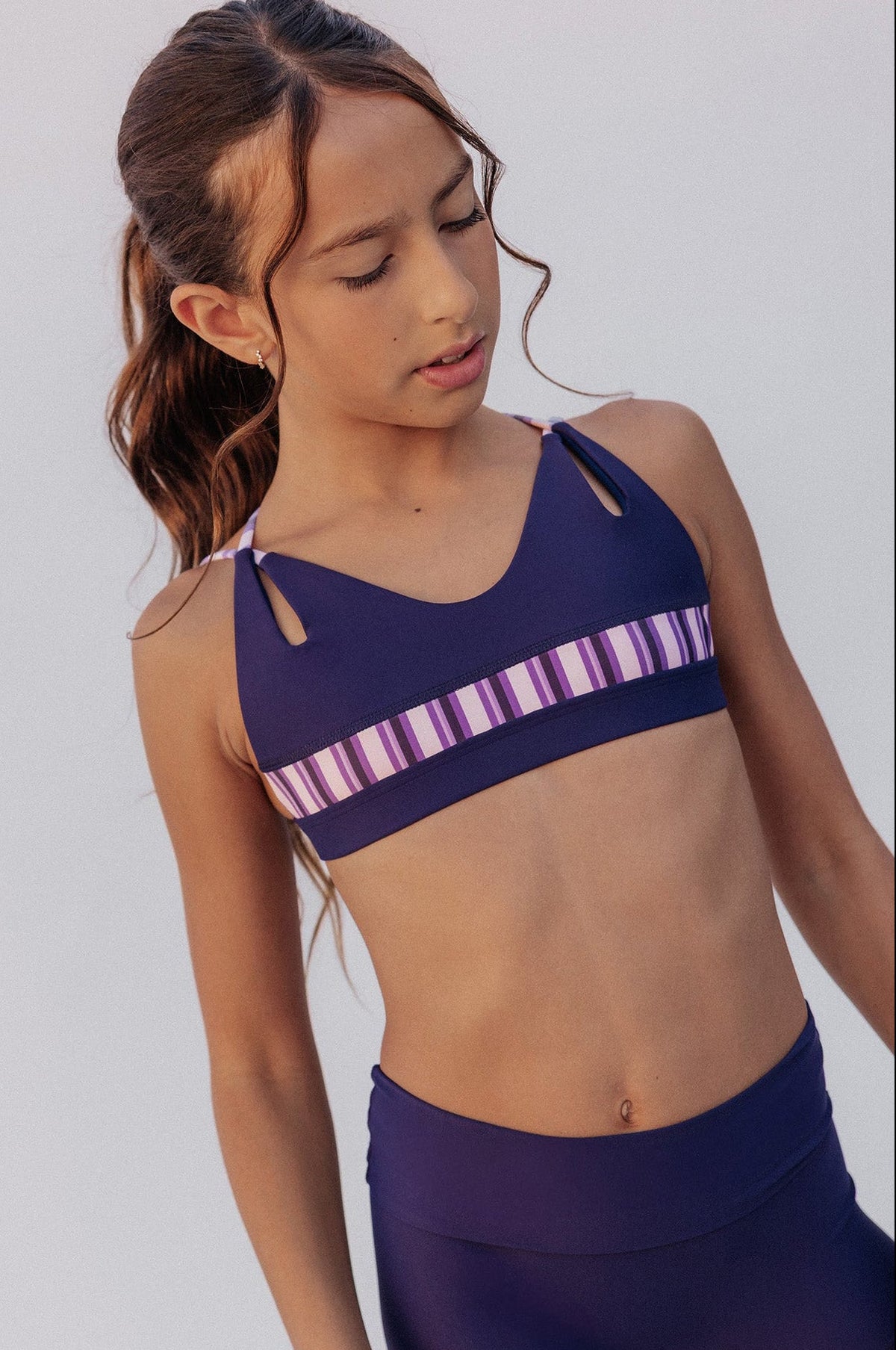 kids, A young girl modeling a two-piece athletic set consisting of a navy blue sports bra with a purple and white striped band, and matching purple shorts, against a light gray studio background.
