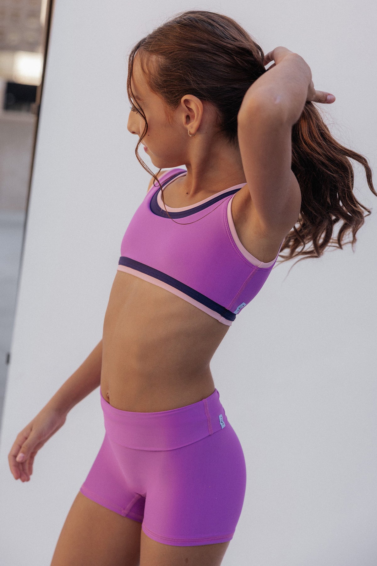 kids, wearing a matching purple athletic set consisting of a sports bra and high-waisted shorts, shown from a side profile against a white studio background.