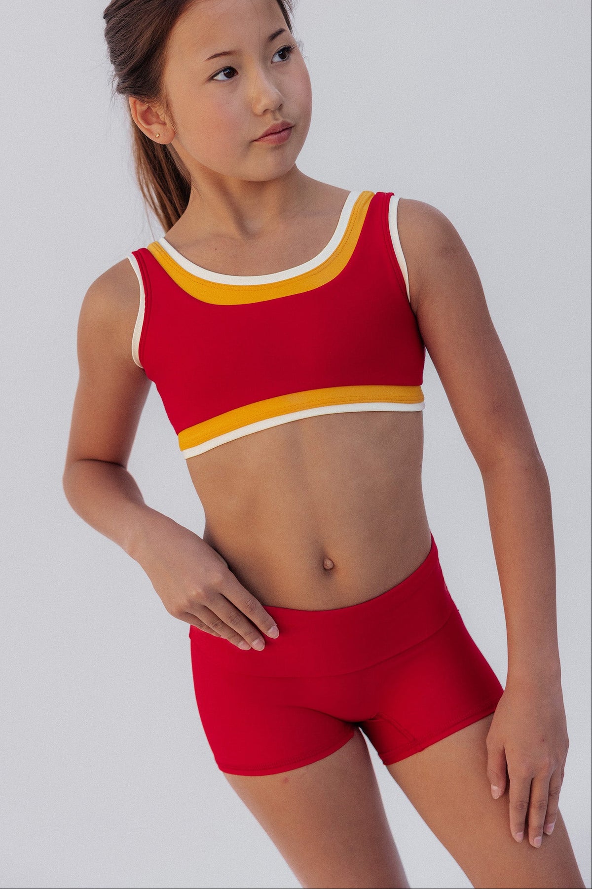 kids, A young girl modeling a red two-piece athletic set consisting of a scoop-neck crop top with yellow and white trim, and matching red athletic shorts, against a light gray studio background