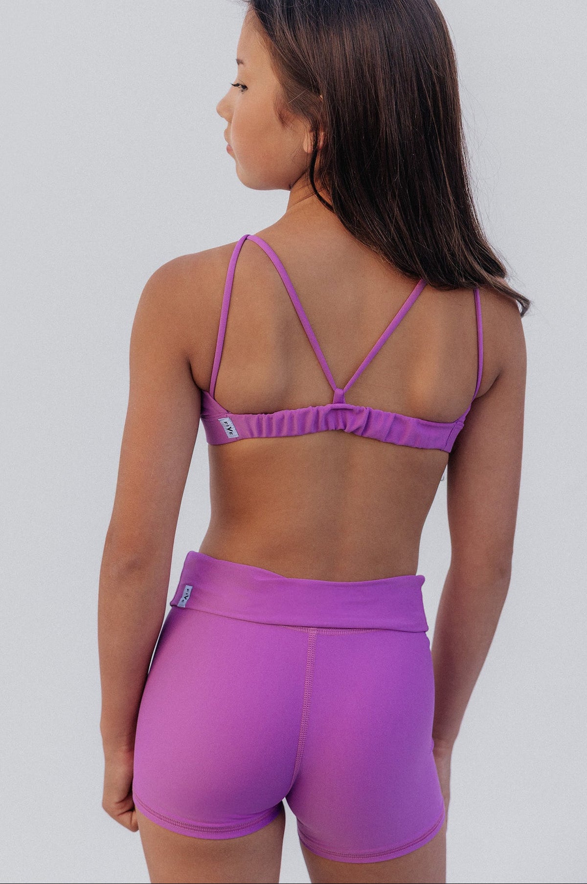 kids, A young girl viewed from behind wearing a matching purple athletic set, featuring a sports bra with crisscross straps and high-waisted shorts, set against a plain light gray background.
