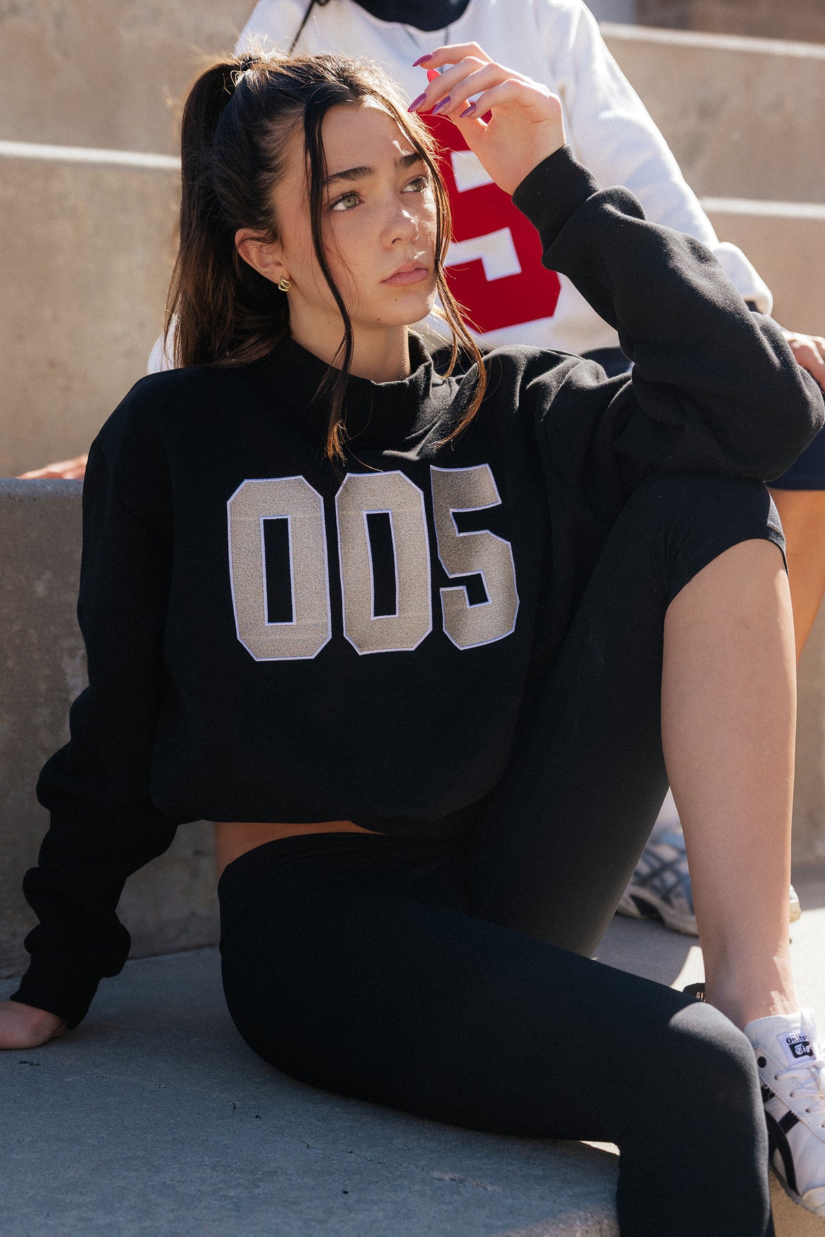 kids, A girl sitting outdoors on concrete steps wearing a black cropped sweatshirt with large silver varsity-style '005' lettering on the front, paired with black leggings.