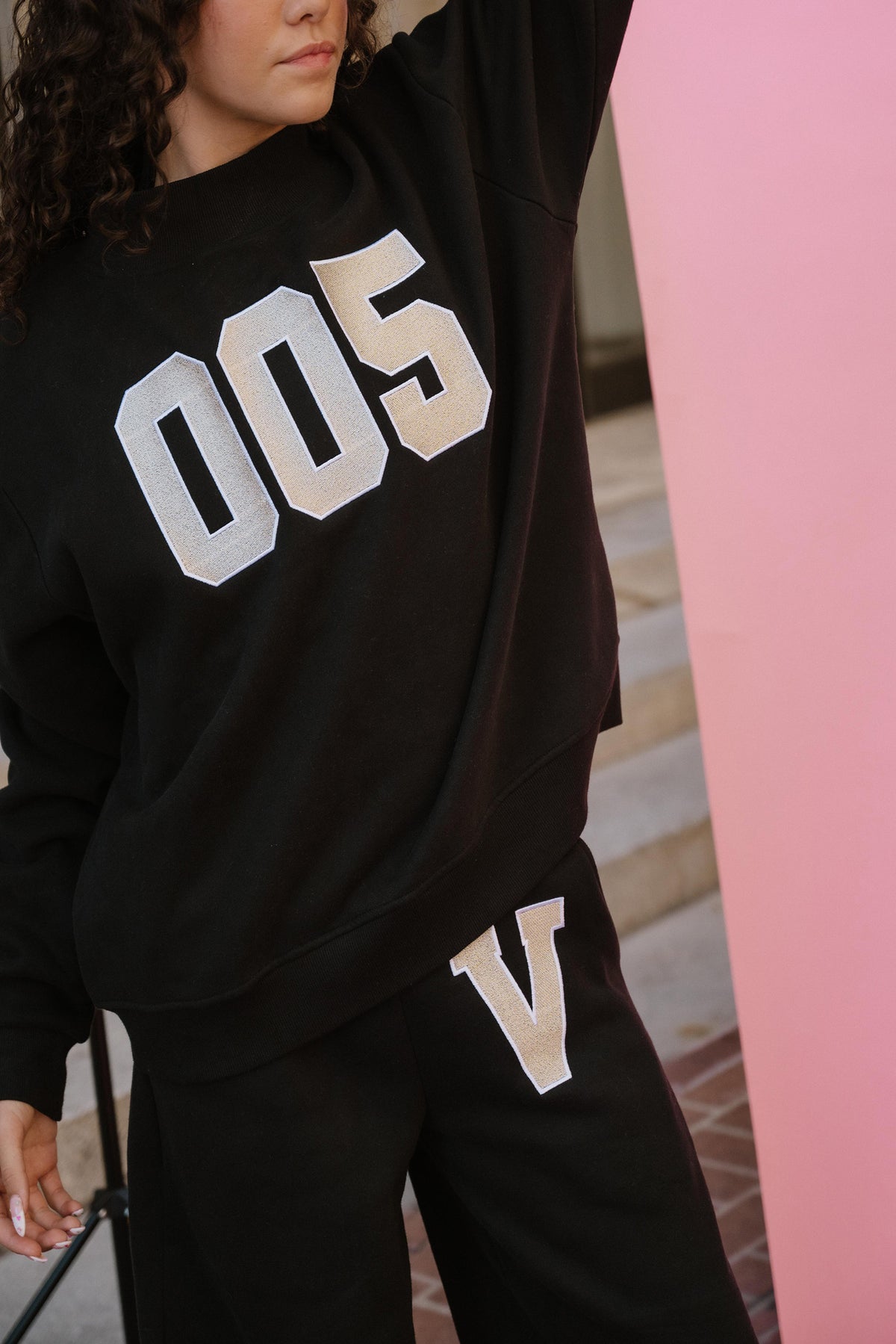 teen, A girl wearing a black sweatshirt and sweatpants set with large metallic silver lettering, standing indoors next to a pink wall. The sweatshirt shows the number '005' and the pants show a large 'V'.